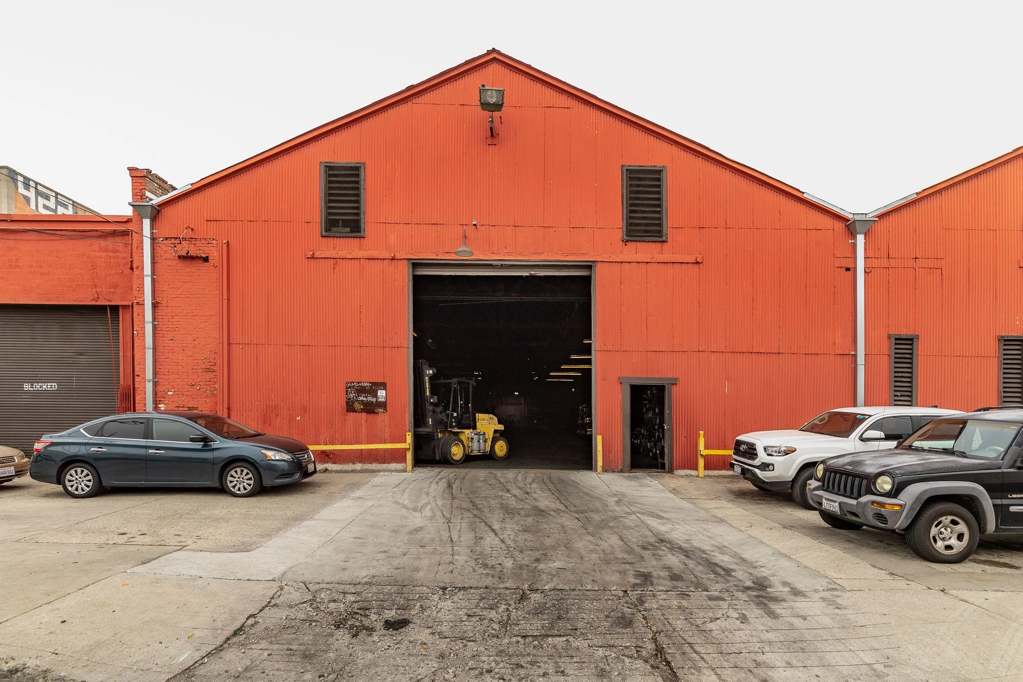 Large red industrial warehouse with an open garage door, cars parked in front, and a forklift inside.