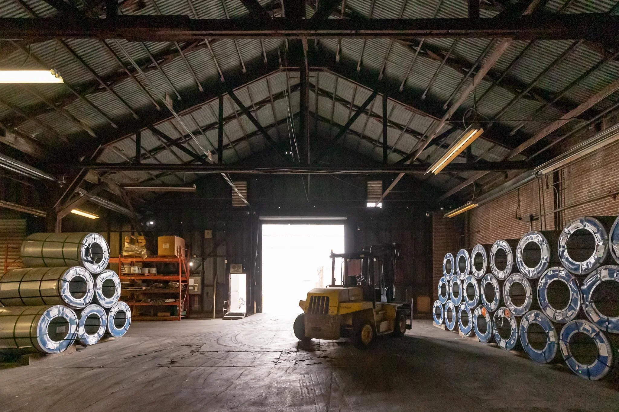 A warehouse interior with a forklift and metal rolls stacked on both sides, open door letting in bright light.