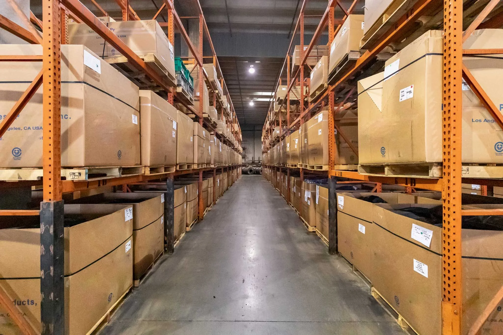 A warehouse aisle with tall orange metal shelves filled with large cardboard boxes and pallets, extending into the distance under bright overhead lighting.