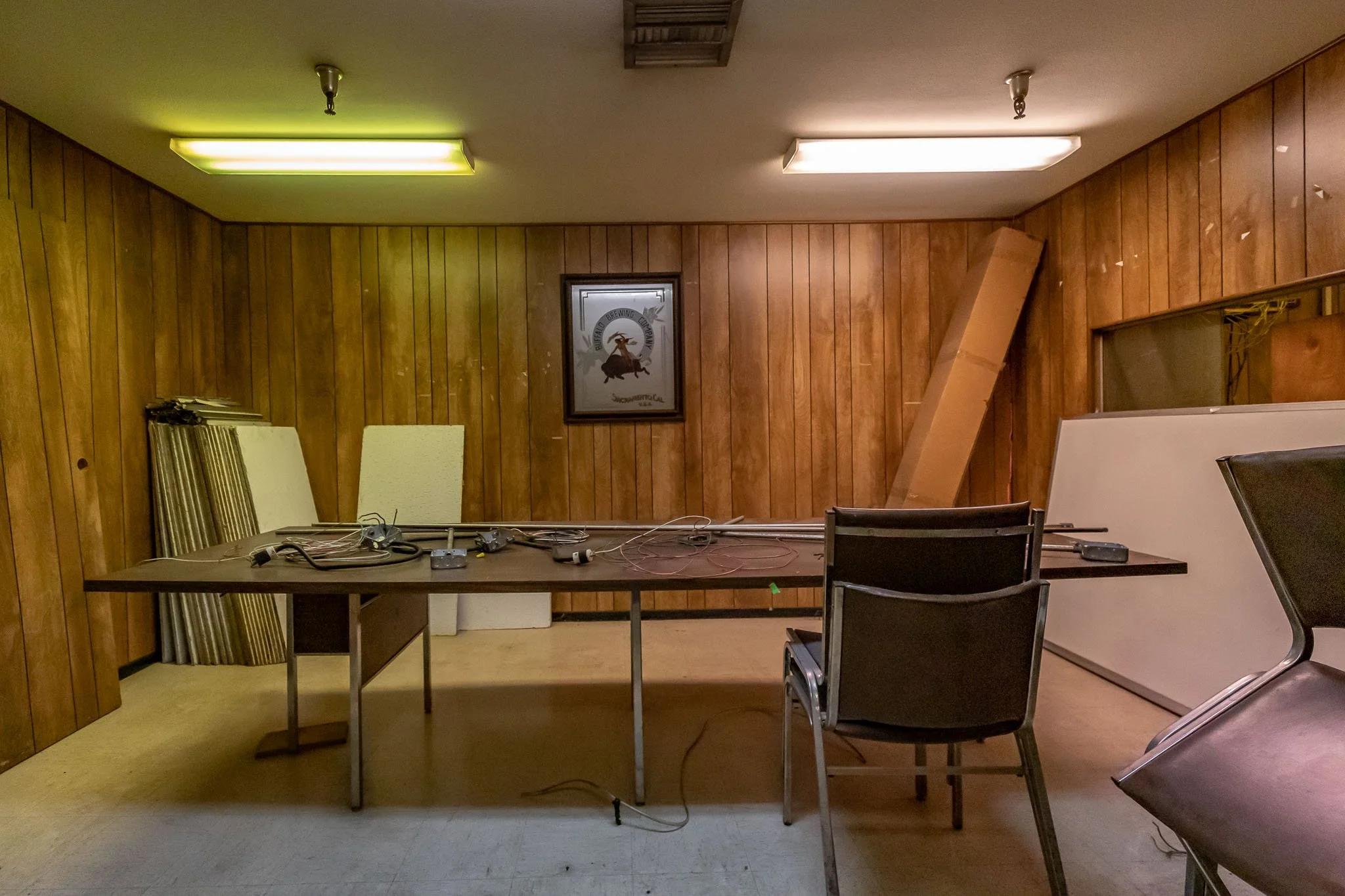 Empty office or conference room with wooden panel walls, a table with electronic equipment, chairs, and some tall panels or boards leaning against the wall.