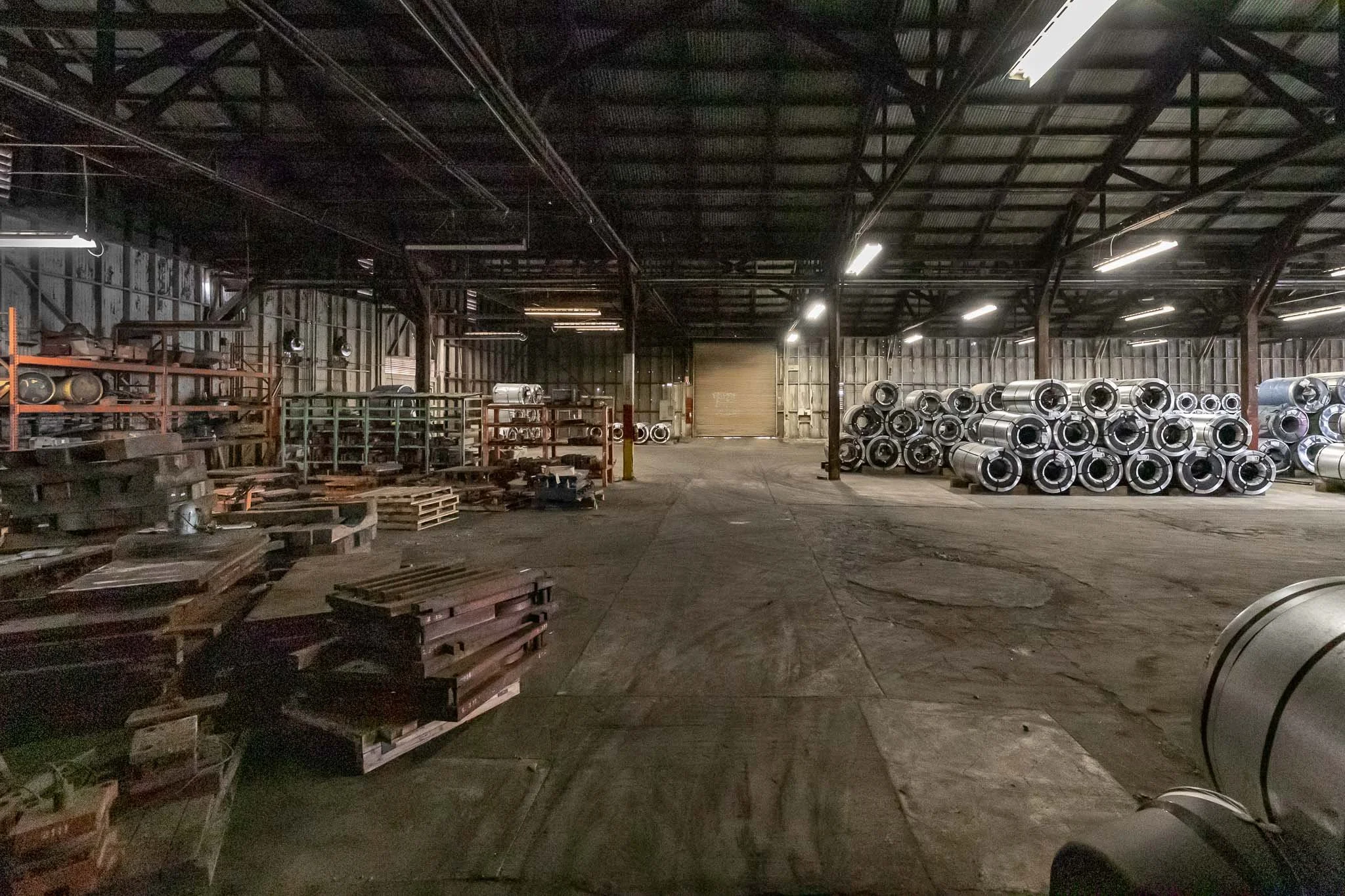 Interior of a large industrial warehouse with metal storage racks, pallets, and cylindrical metal rolls stacked on the right, with a concrete floor and metal walls, illuminated by overhead lighting.