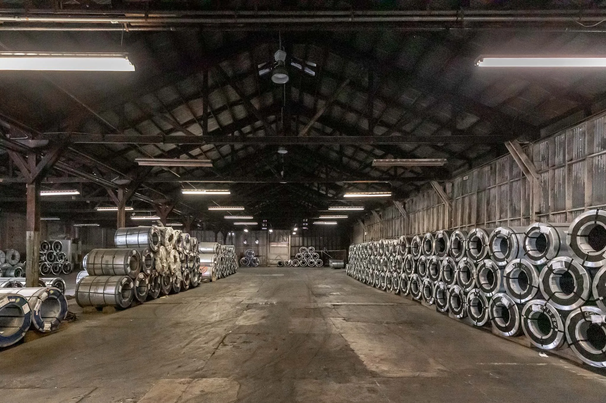 Steel coils stored in an industrial warehouse with a high vaulted ceiling and exposed wooden beams.