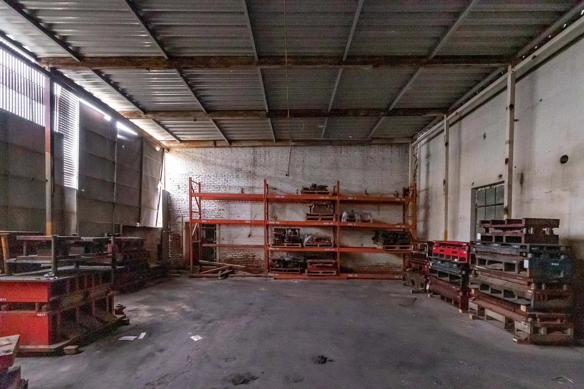 Empty industrial warehouse with metal shelving, some stacked pallets, and a brick wall, illuminated by natural light.