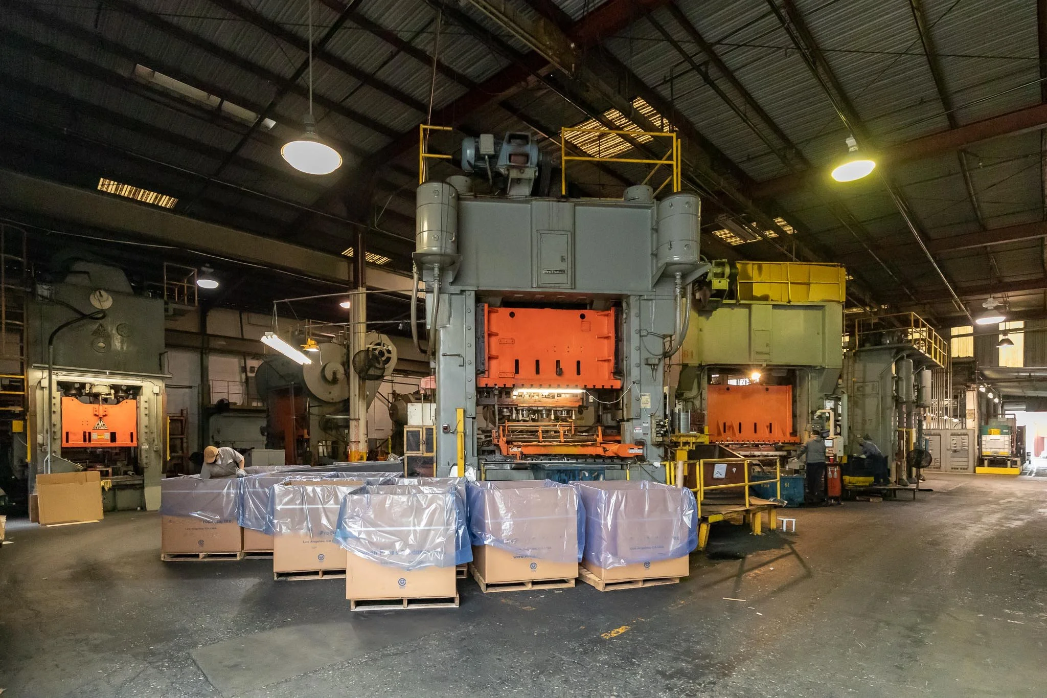 An industrial factory interior with large machinery, workers, and pallets covered with plastic in the foreground.