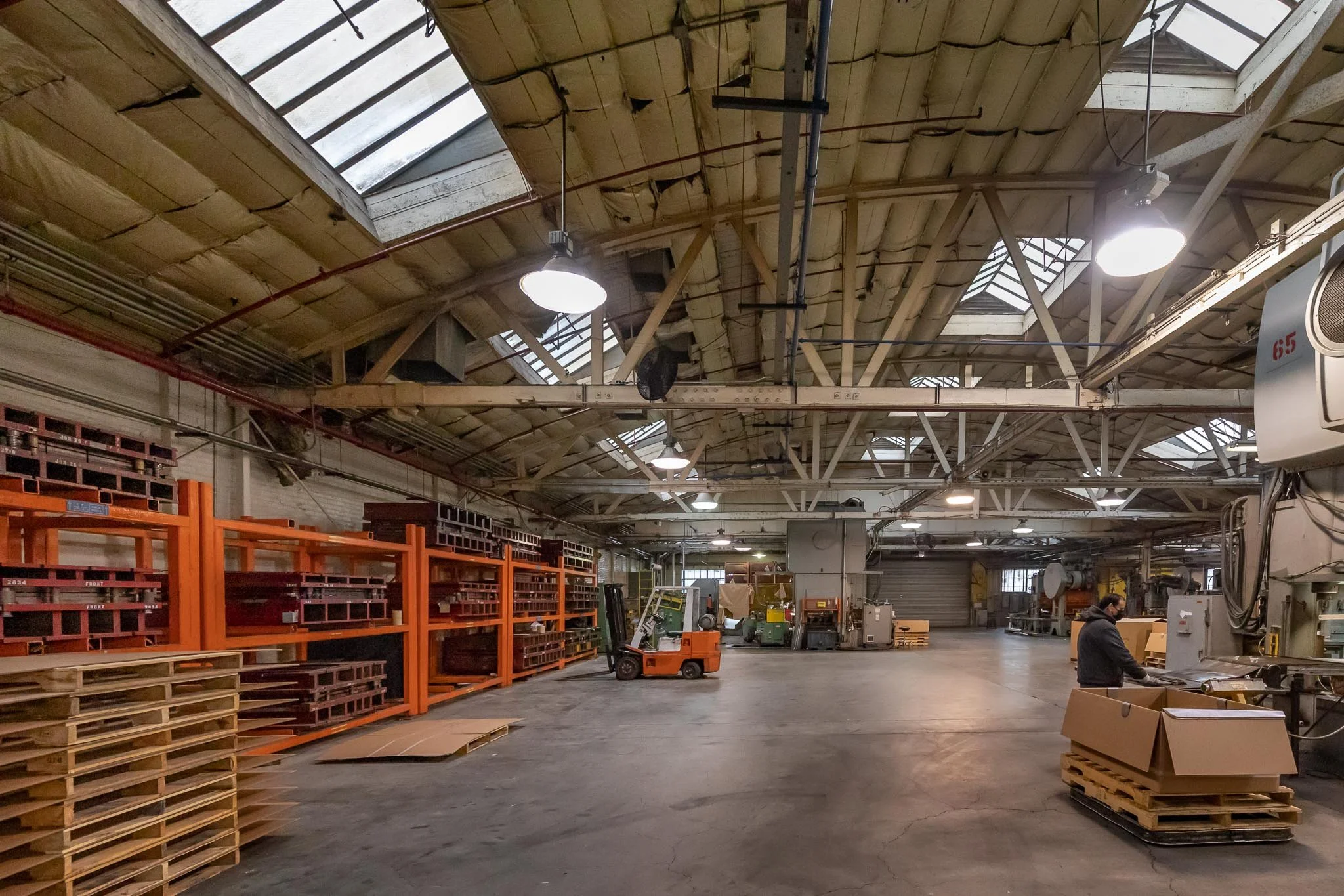Interior view of a large industrial warehouse with stacks of wooden pallets, orange shelving units, manufacturing equipment, and a worker operating machinery.