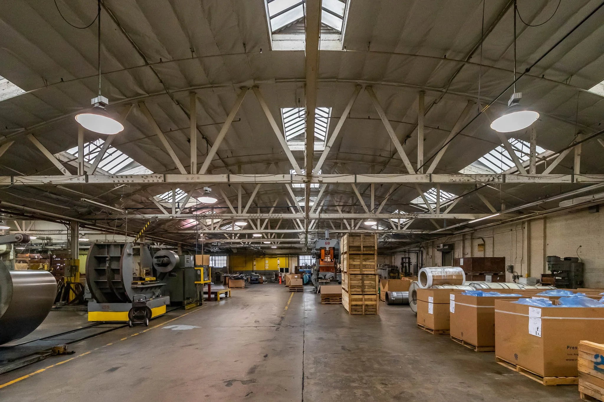 Empty industrial warehouse with large machinery, wooden pallets, and metal rolls, illuminated by overhead lights and skylights.