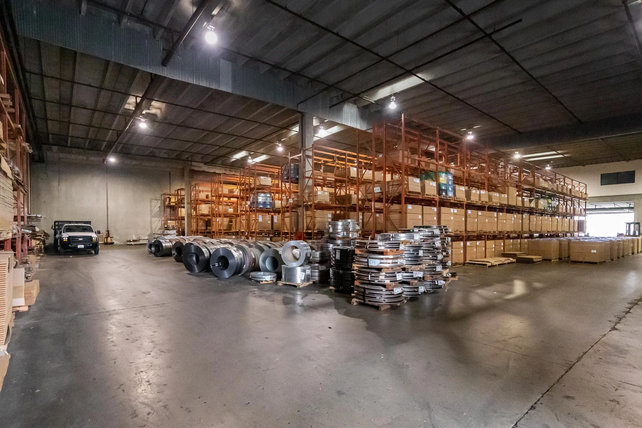 A large warehouse with metal shelves filled with boxes and industrial equipment, a white truck parked on the left side, and metal coils and stacks of flat metal pieces on the floor.