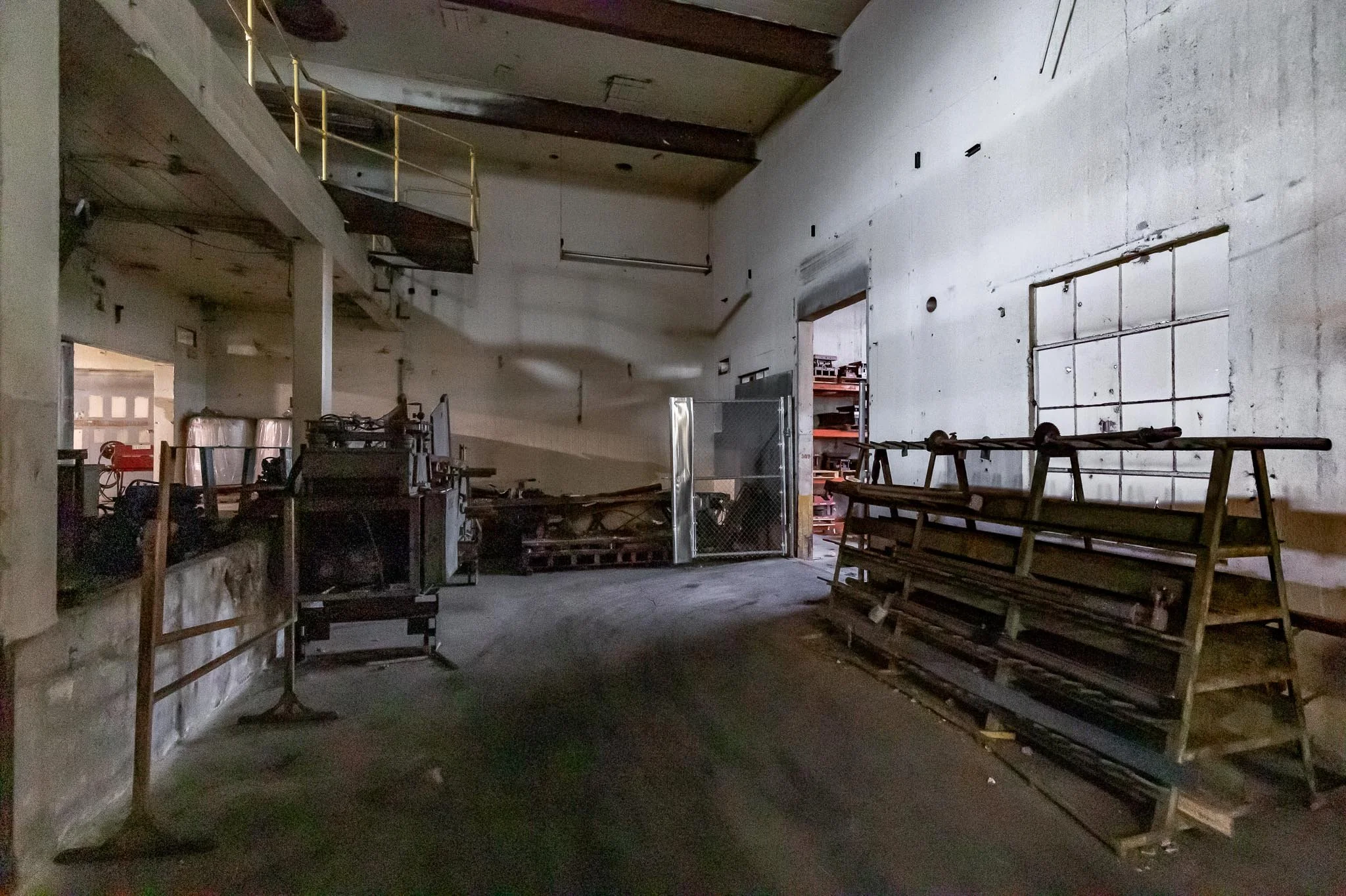 Empty industrial warehouse with scattered equipment, abandoned shelves, and a closed grated door, dimly lit with concrete walls and floor.