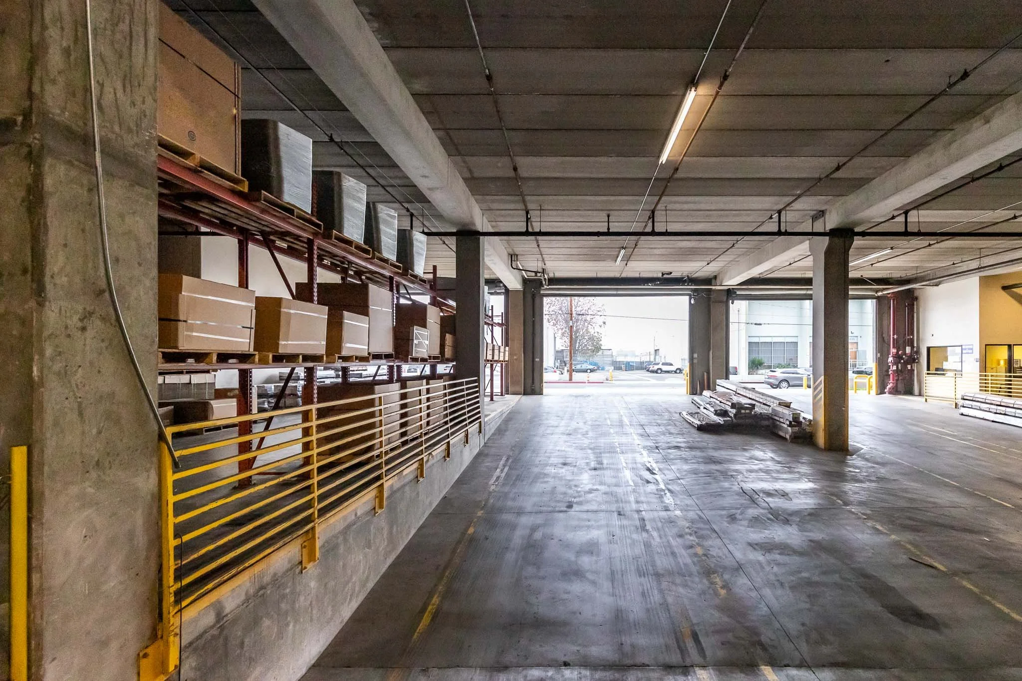 Empty warehouse loading dock with metal shelving, boxes, and construction materials, open to outside parking area.