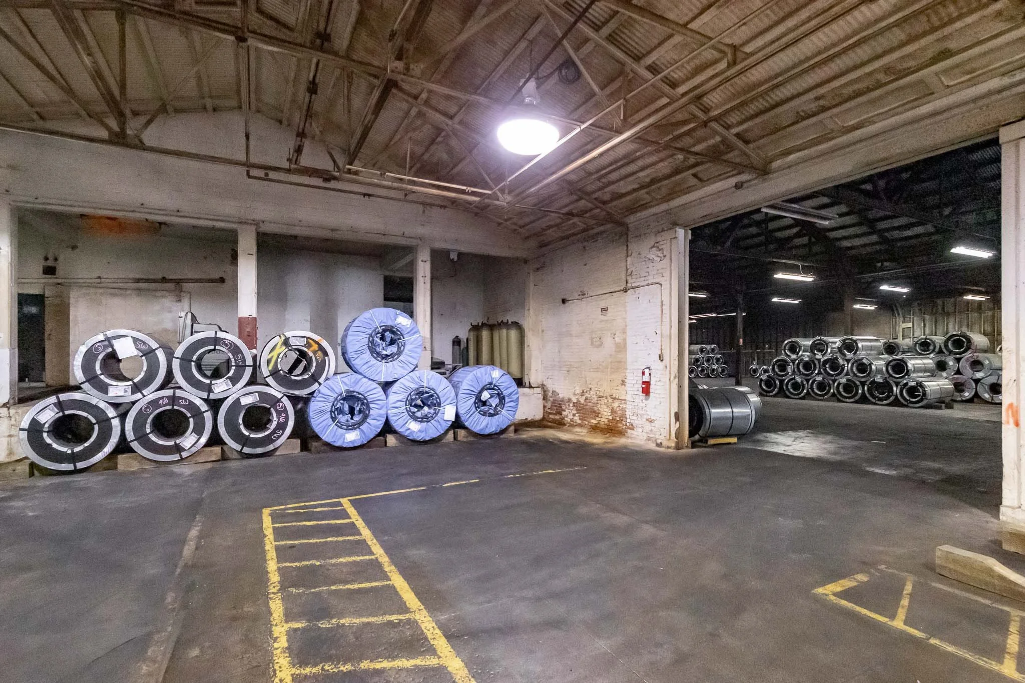 Industrial warehouse with stacked metal rolls and pipes, concrete floors, white brick walls, and overhead lighting.
