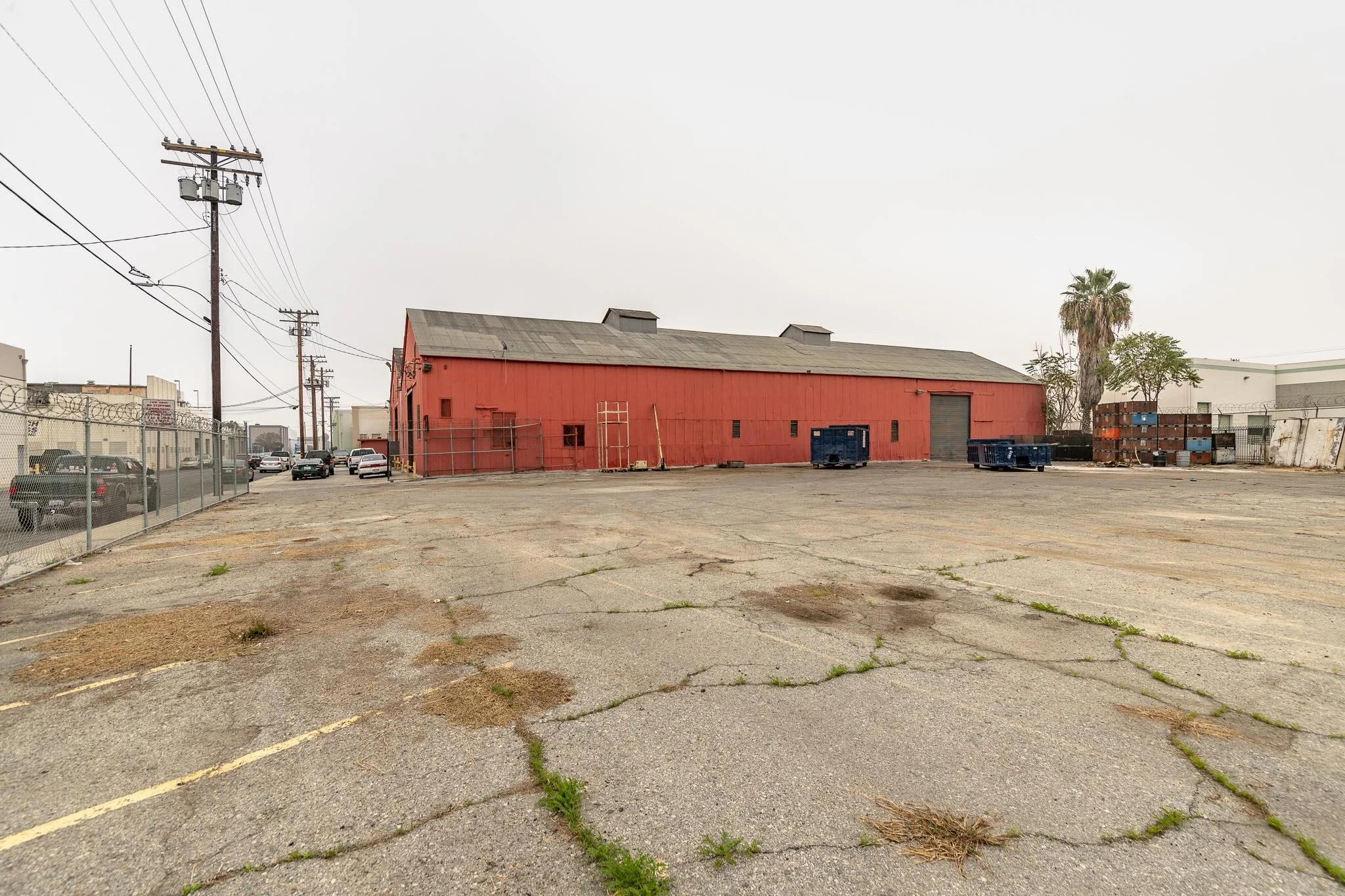 An empty parking lot with cracks and weeds, behind a red barn-like building with a wide door, a few small windows, and a ladder leaning against it. There are some parking cars on the left side and some blue containers and a palm tree on the right sid