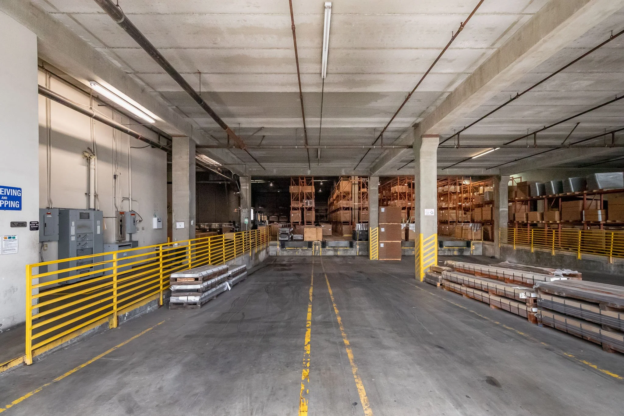 Empty loading dock area in a warehouse with yellow safety barriers, stacked pallets, and shelving units filled with boxes and materials.