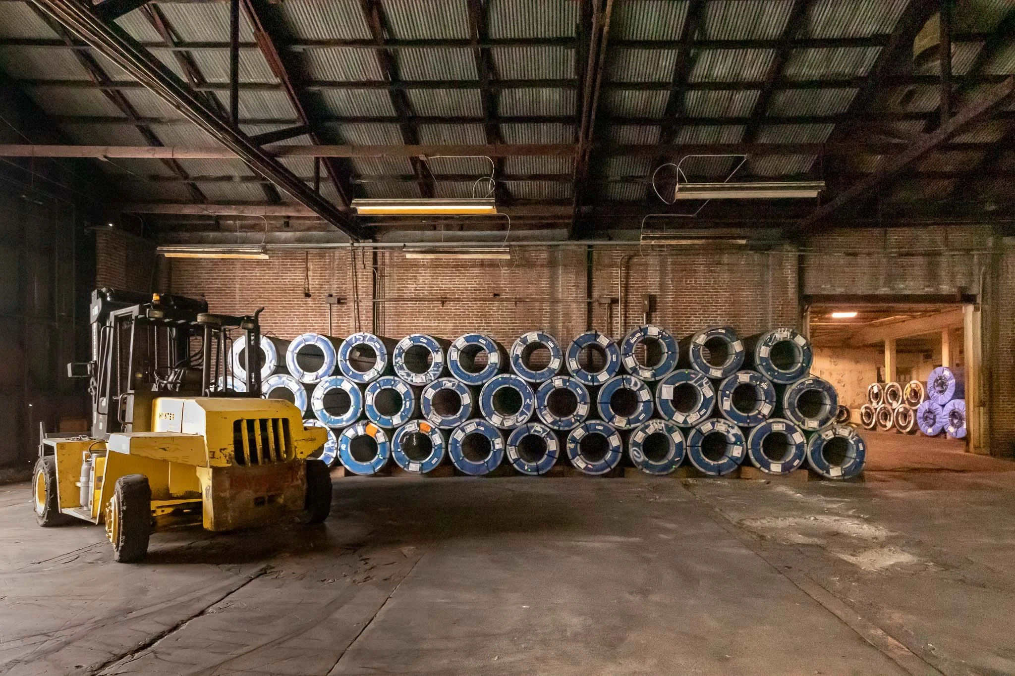 Industrial warehouse with stacks of large blue metal pipes and a yellow forklift in the foreground.
