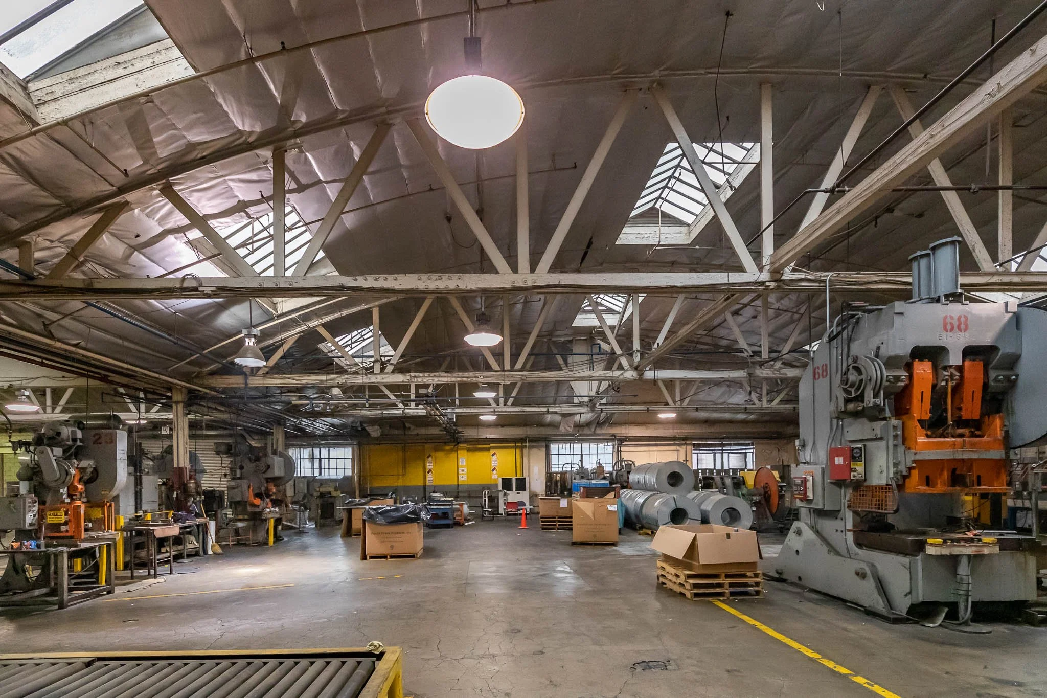 Industrial manufacturing facility with large machines, metal rolls, and workstations in a spacious warehouse with skylights and high ceilings.