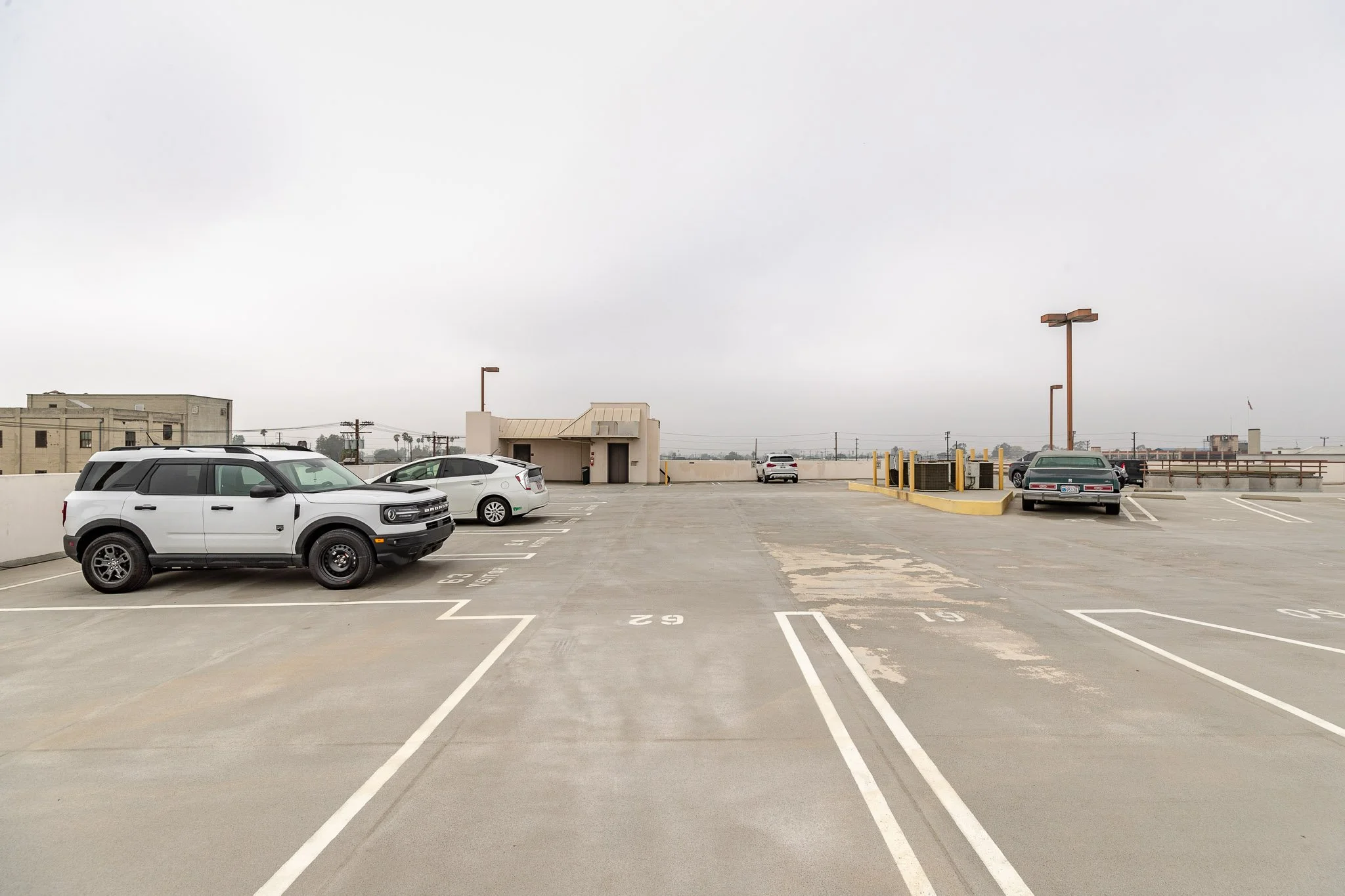 Empty rooftop parking lot with a few cars parked on the sides, overcast sky, city buildings and power lines in the background.