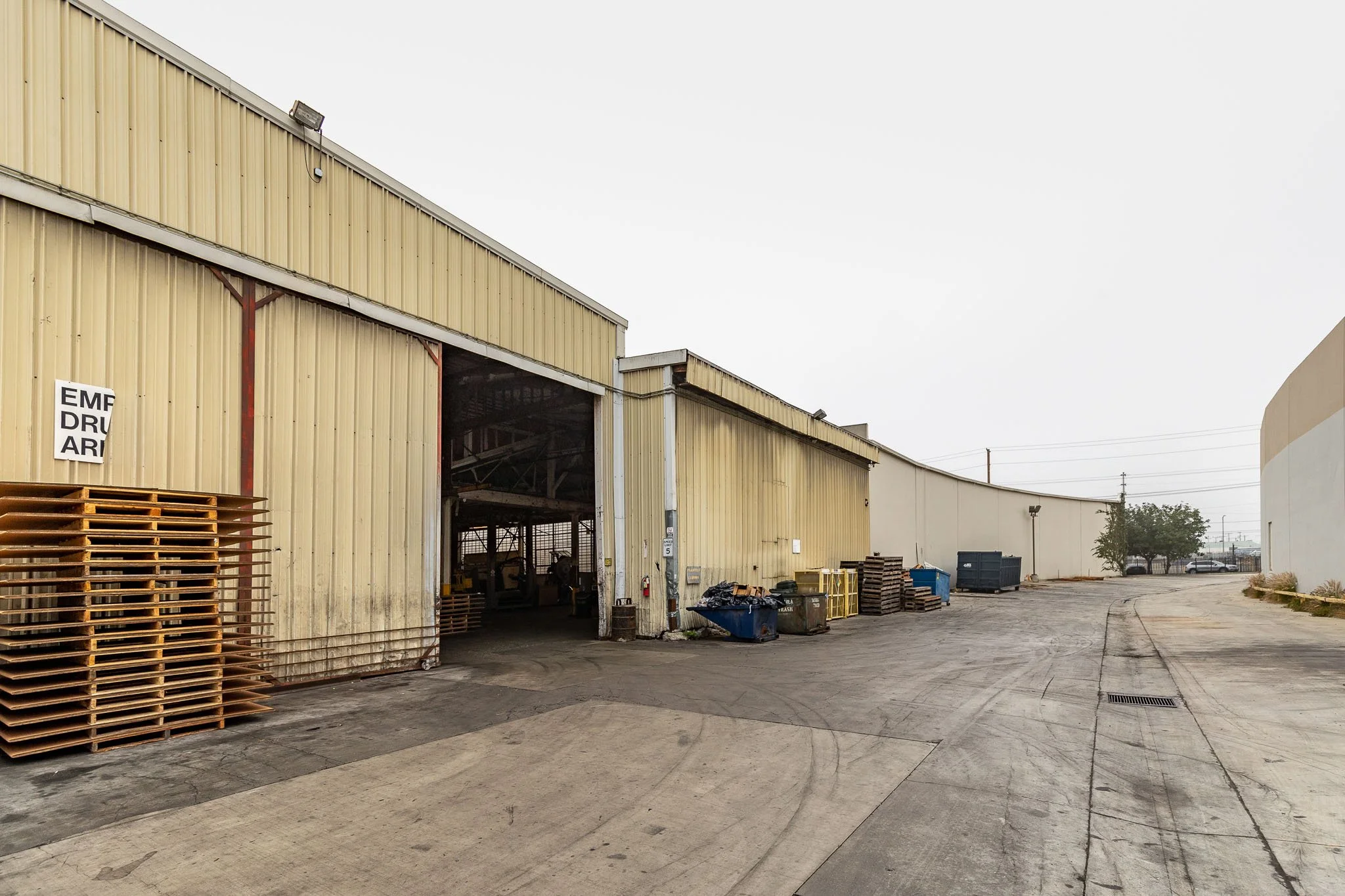 An industrial warehouse with a partially open entrance, surrounded by dumpsters and pallets, in an overcast setting.