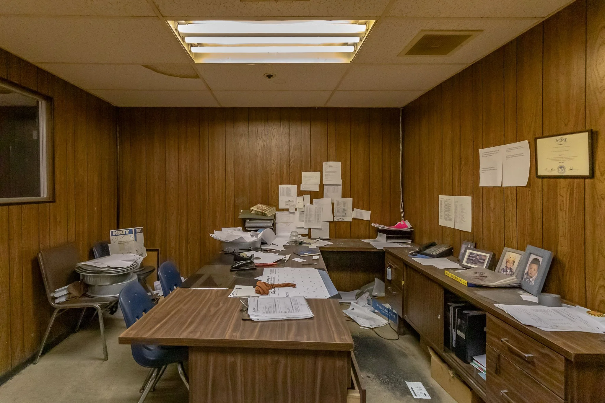  cluttered office with wood paneled walls, disorganized desk, paper stacks, frames with photos, and certificates