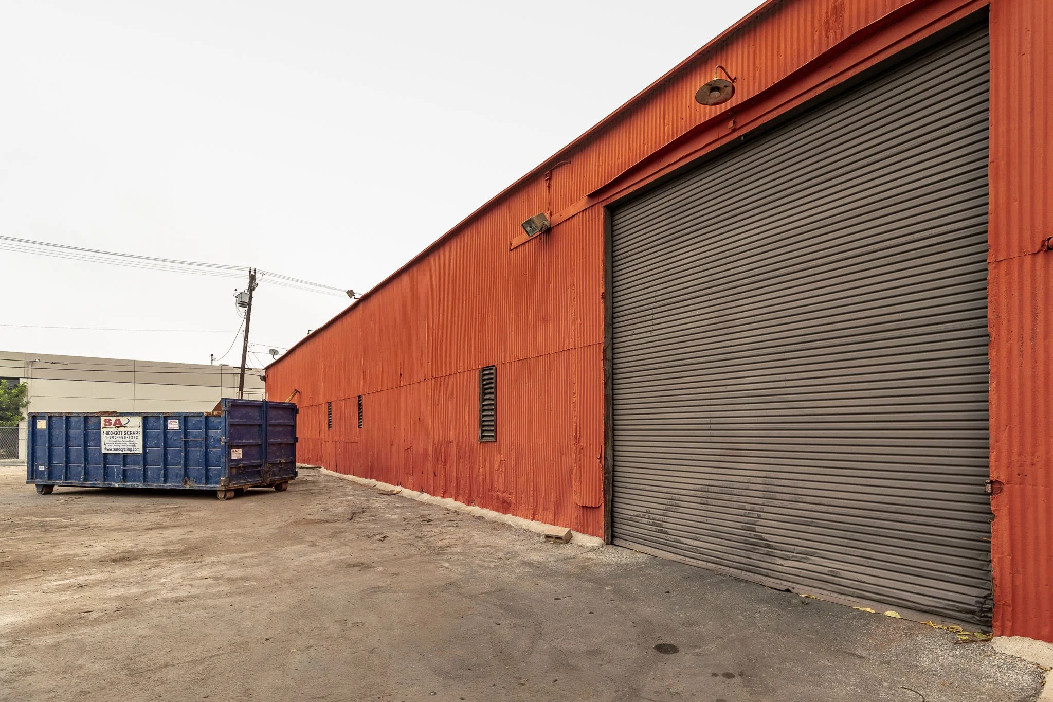 Red industrial building with a large gray rolling door and two small vents, a blue dumpster nearby, and utility poles in the background.