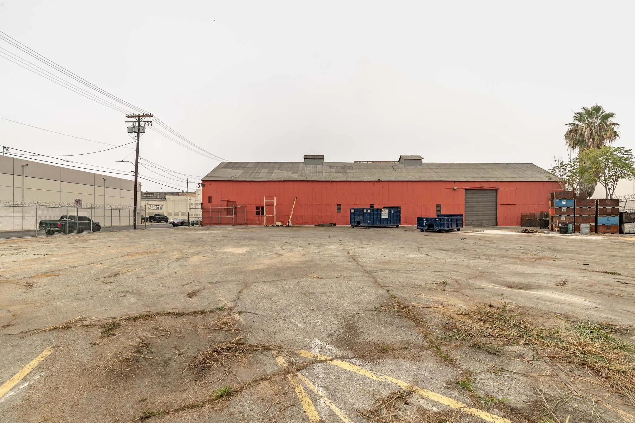 An empty parking lot in front of a red warehouse building with stacked blue and brown pallets, a large closed garage door, and a few scattered trees. Overcast sky.