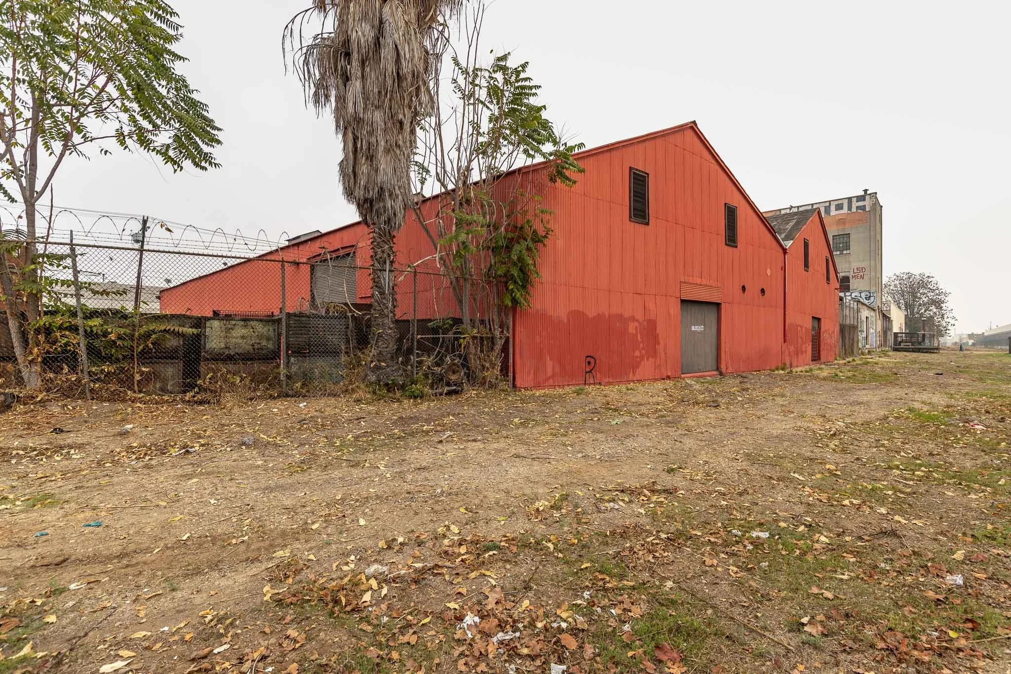 A large red industrial building with a metal garage door and small windows, surrounded by dirt and scattered leaves, with a barbed wire fence and trees nearby, under a foggy sky.