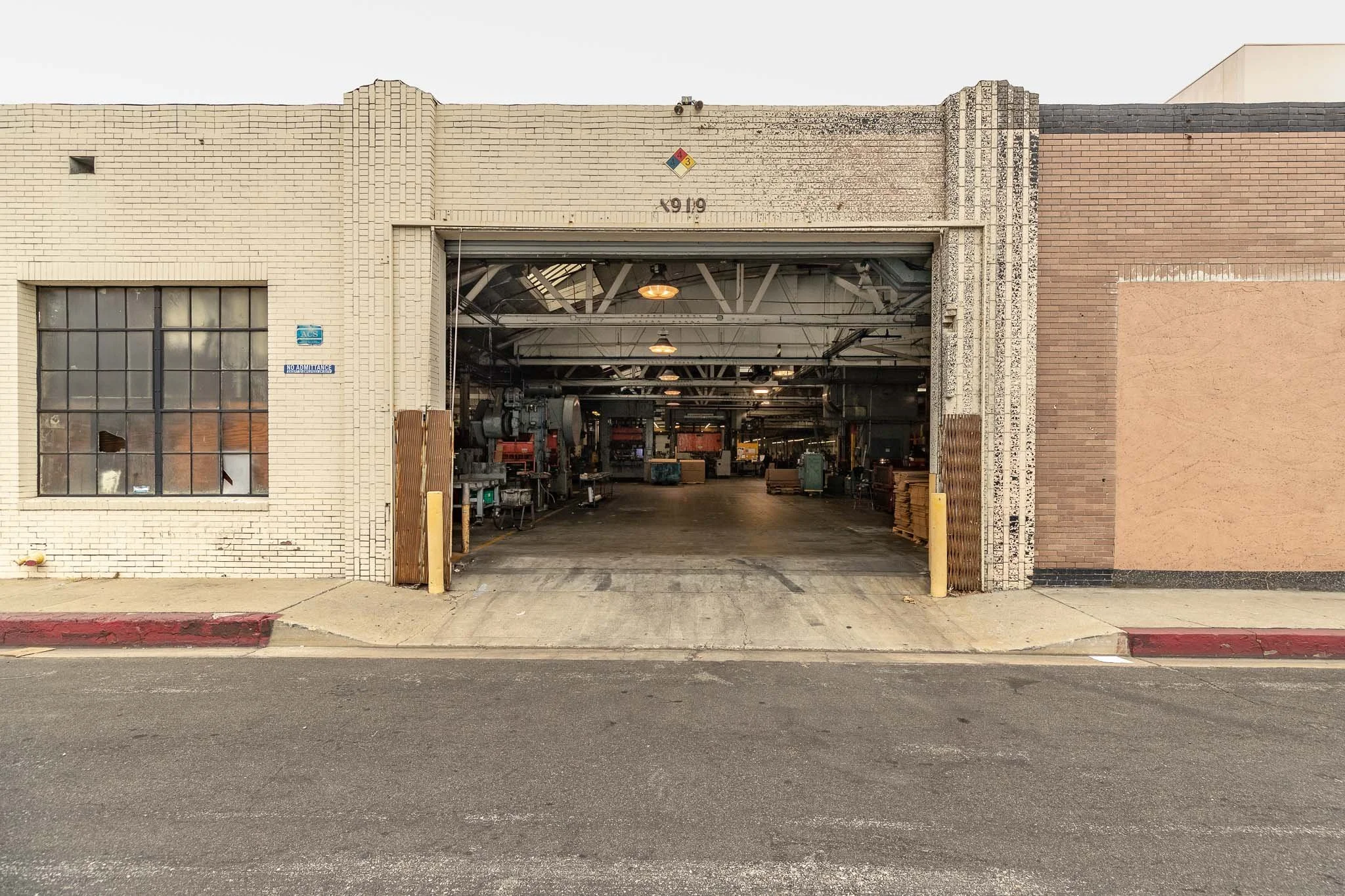 View of a large warehouse or industrial building with a wide open garage door revealing the interior filled with machinery and equipment, brick walls, and a sidewalk in front.