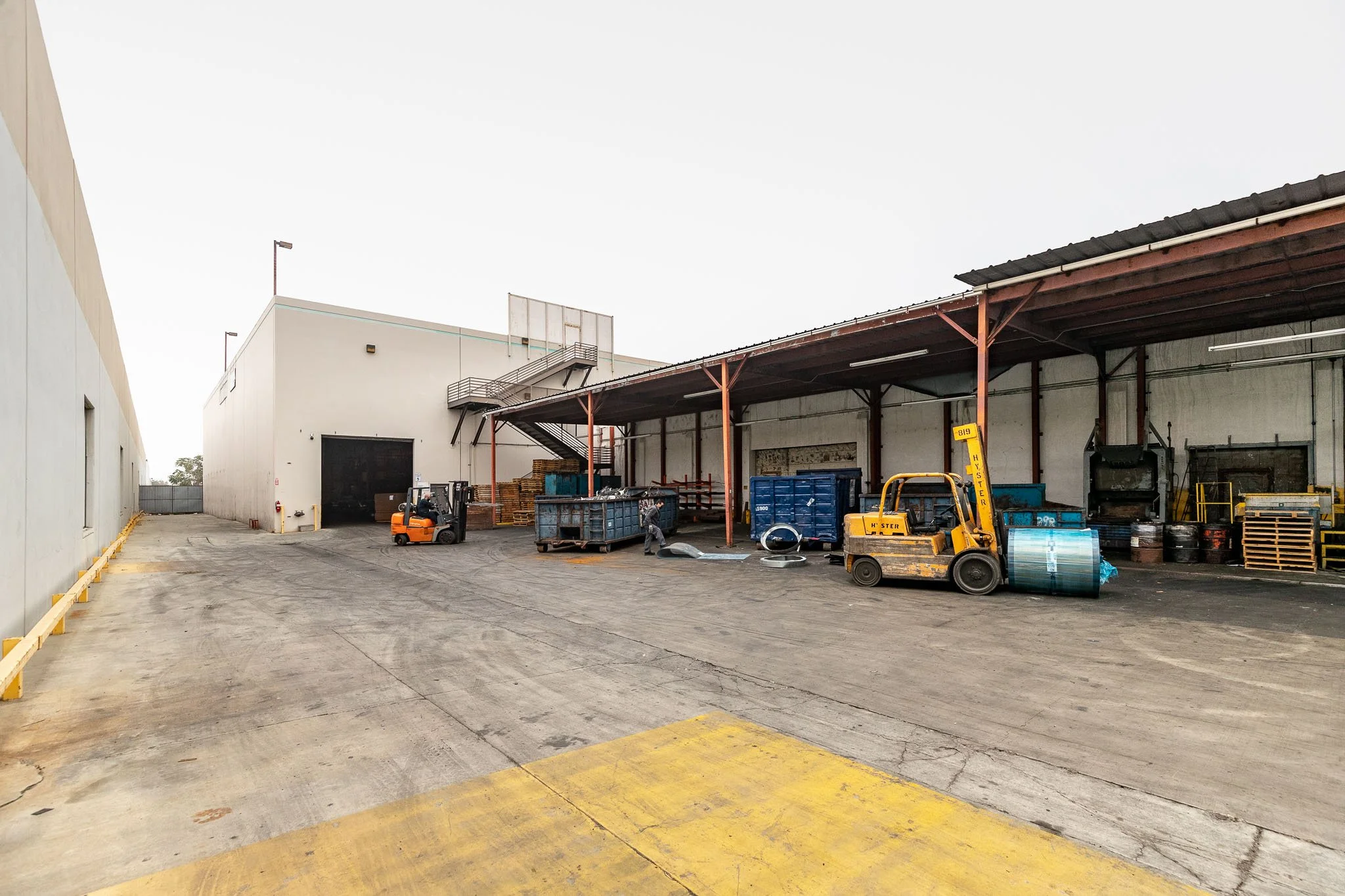 An industrial loading area with forklifts, pallets, and storage containers under a covered structure attached to a large white warehouse.