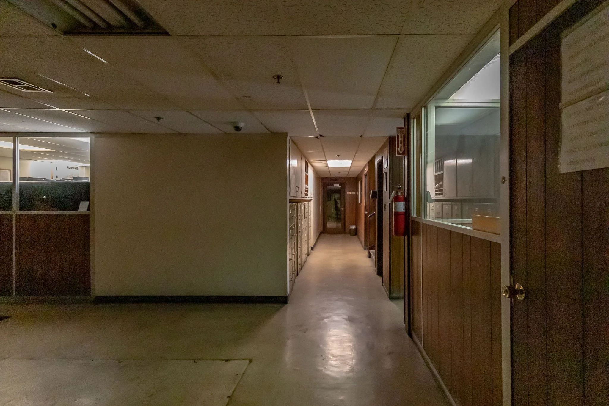 Empty hallway with wood-paneled walls, mailboxes on the left, and a fire extinguisher mounted on the wall on the right, lit by ceiling lights.