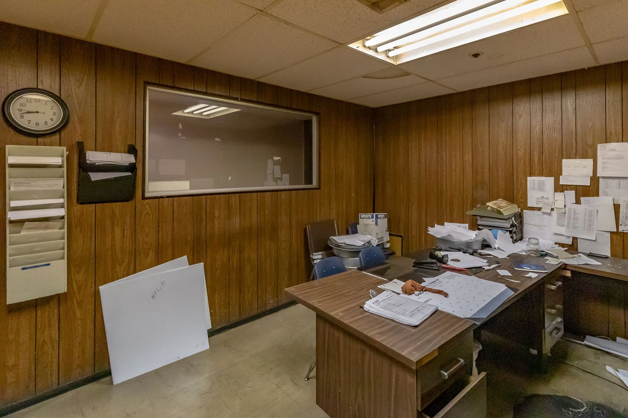 An office room with wood-paneled walls, cluttered desk with papers, folders, and office supplies, and a window with a reflecting light. The room appears messy and disorganized.