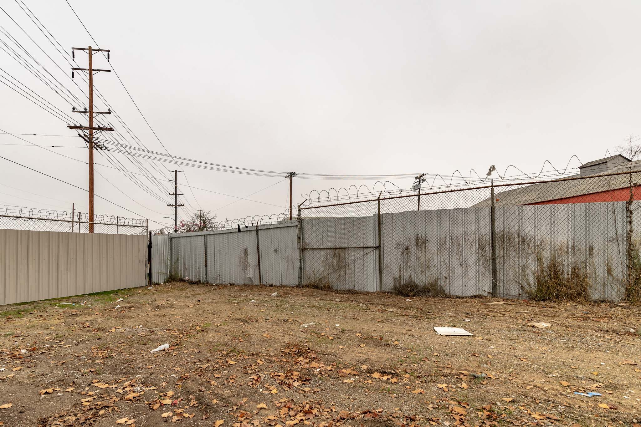 Barbed wire-topped chain-link fence with a closed metal gate, on a dirt lot with scattered leaves and trash, under a cloudy sky and power lines overhead.