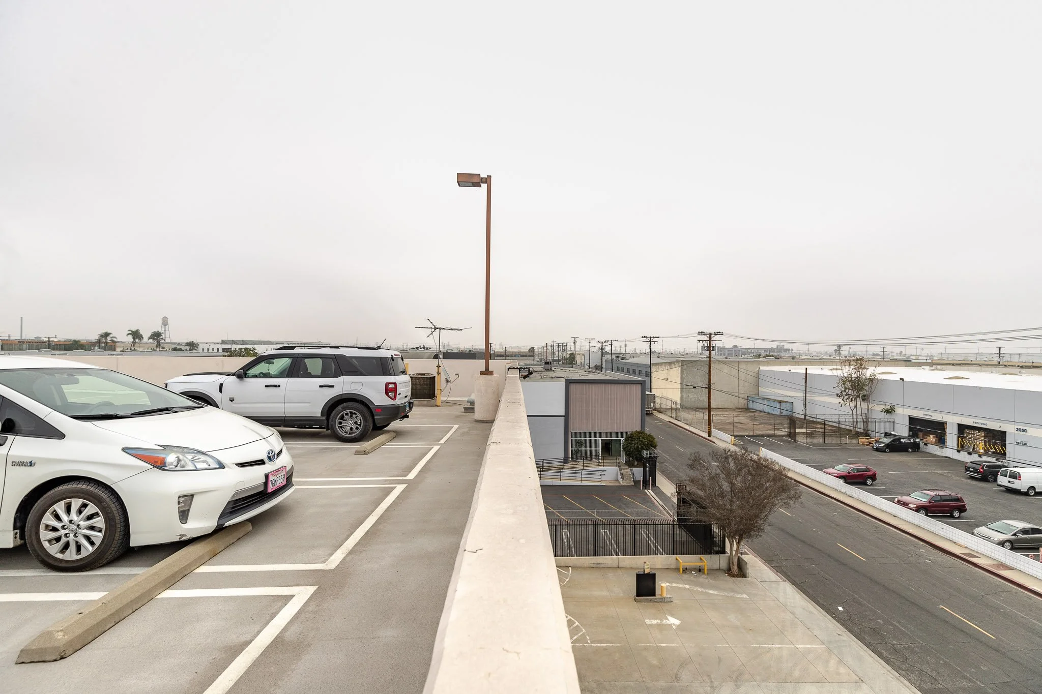 View of an empty rooftop parking lot with a few parked cars, a street below, industrial buildings, utility poles, and an overcast sky.
