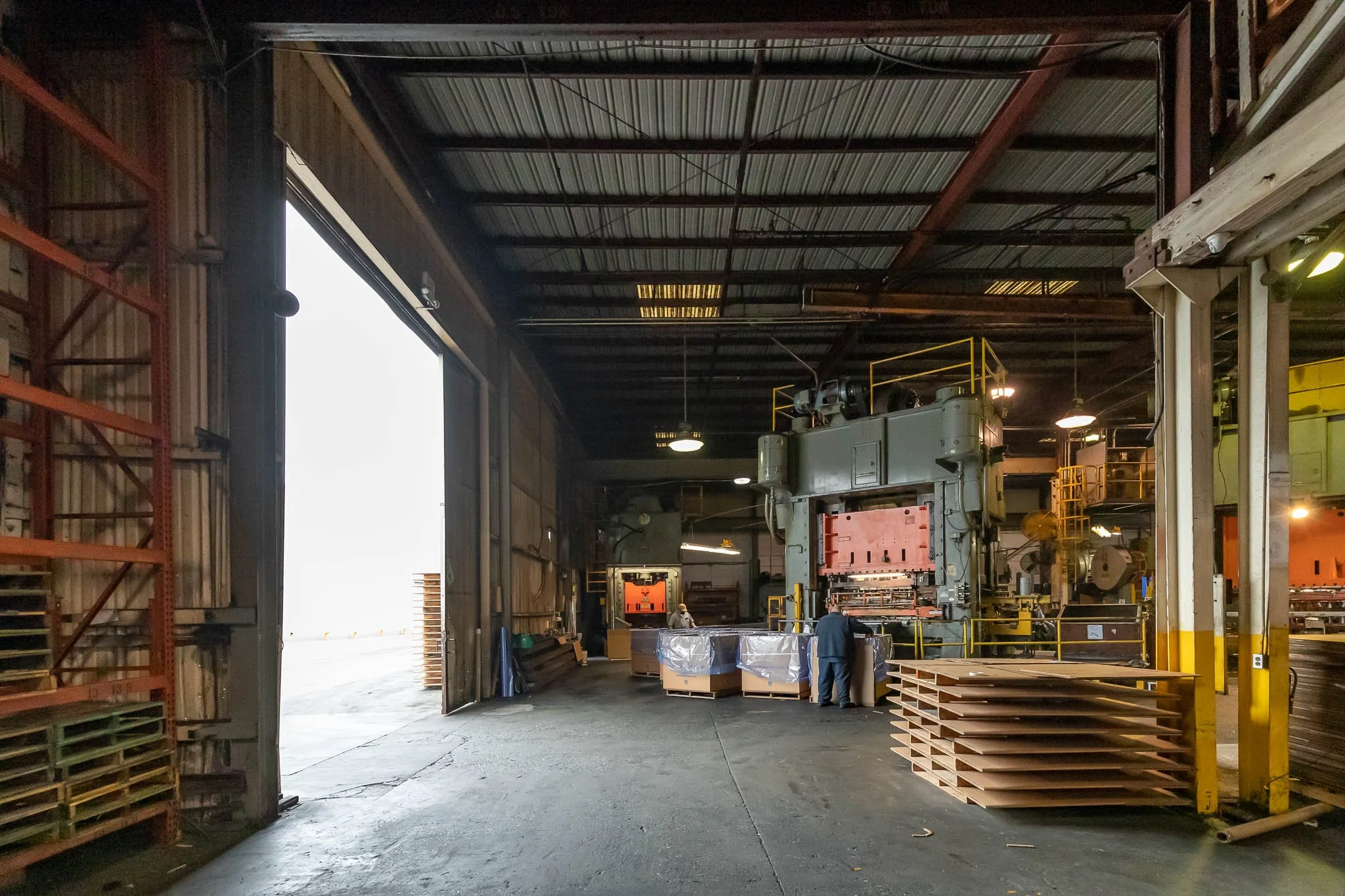 Inside a large industrial warehouse with machinery and stacks of materials, partially lit, with an open large door revealing bright light outside.