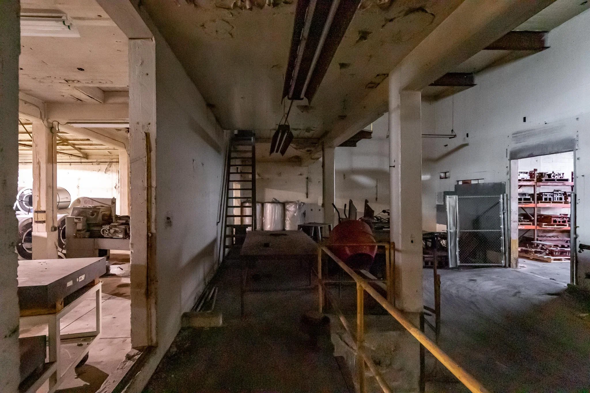 An interior view of an abandoned or unfinished industrial space with clutter and construction tools, including a ladder, a red concrete mixer, and metal shelving.
