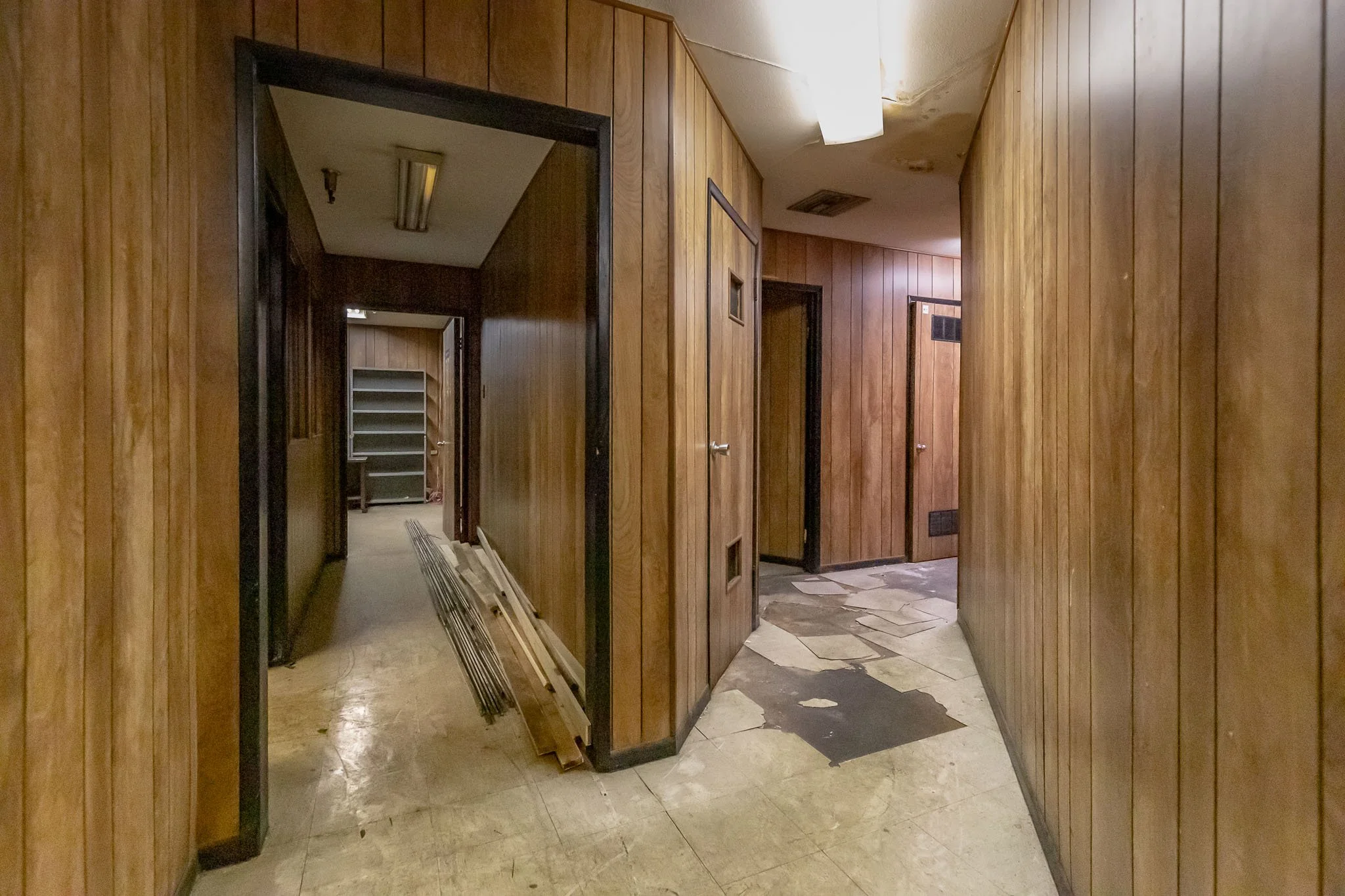 Old hallway with wood-paneled walls, worn flooring, and missing ceiling tiles, leading to a room with shelves and construction materials.