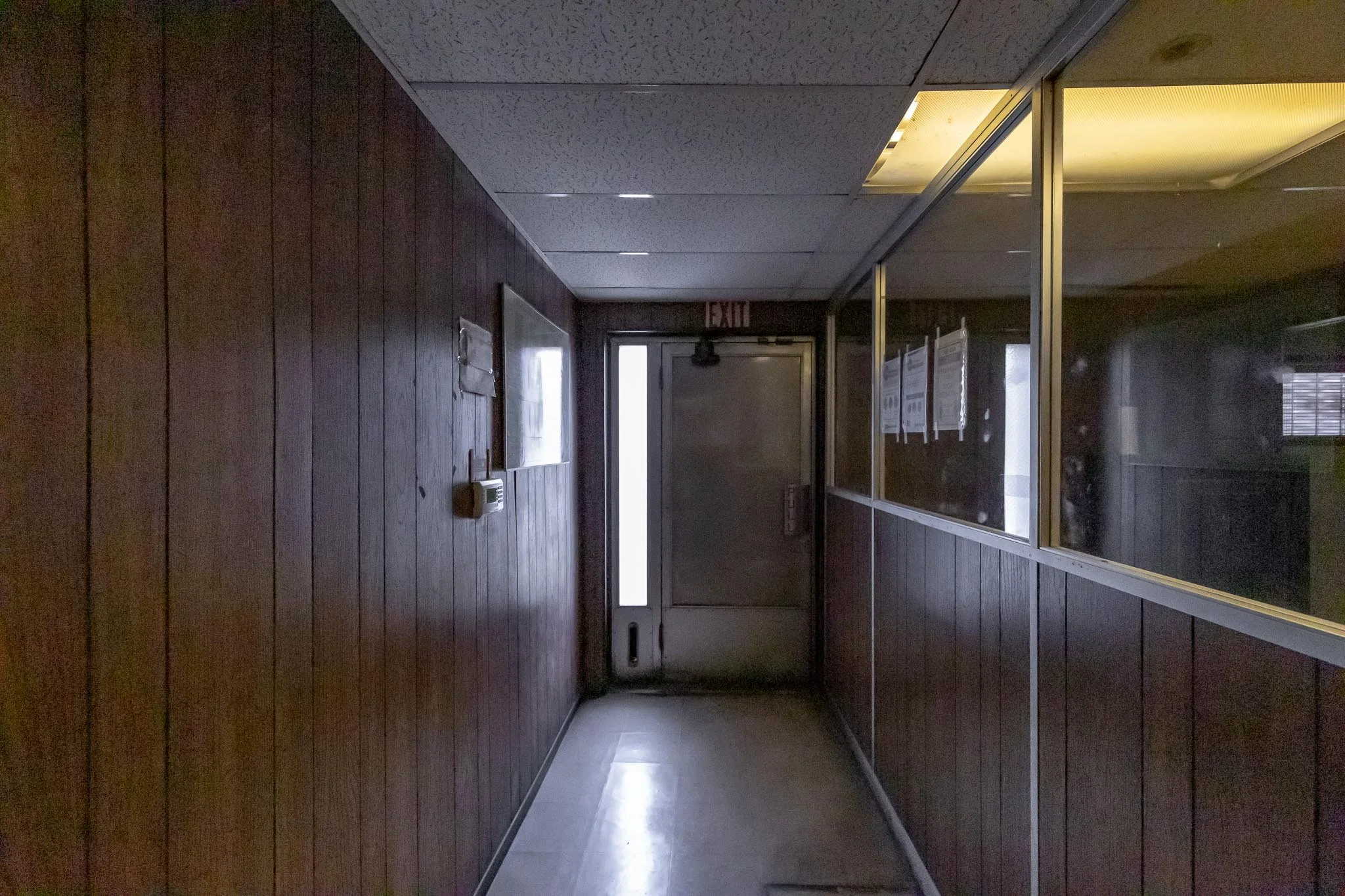 Dark wood-paneled hallway leading to a metal door with an illuminated exit sign above, small window on the door, mirror on the right wall, and notice boards with papers behind glass on the right side.