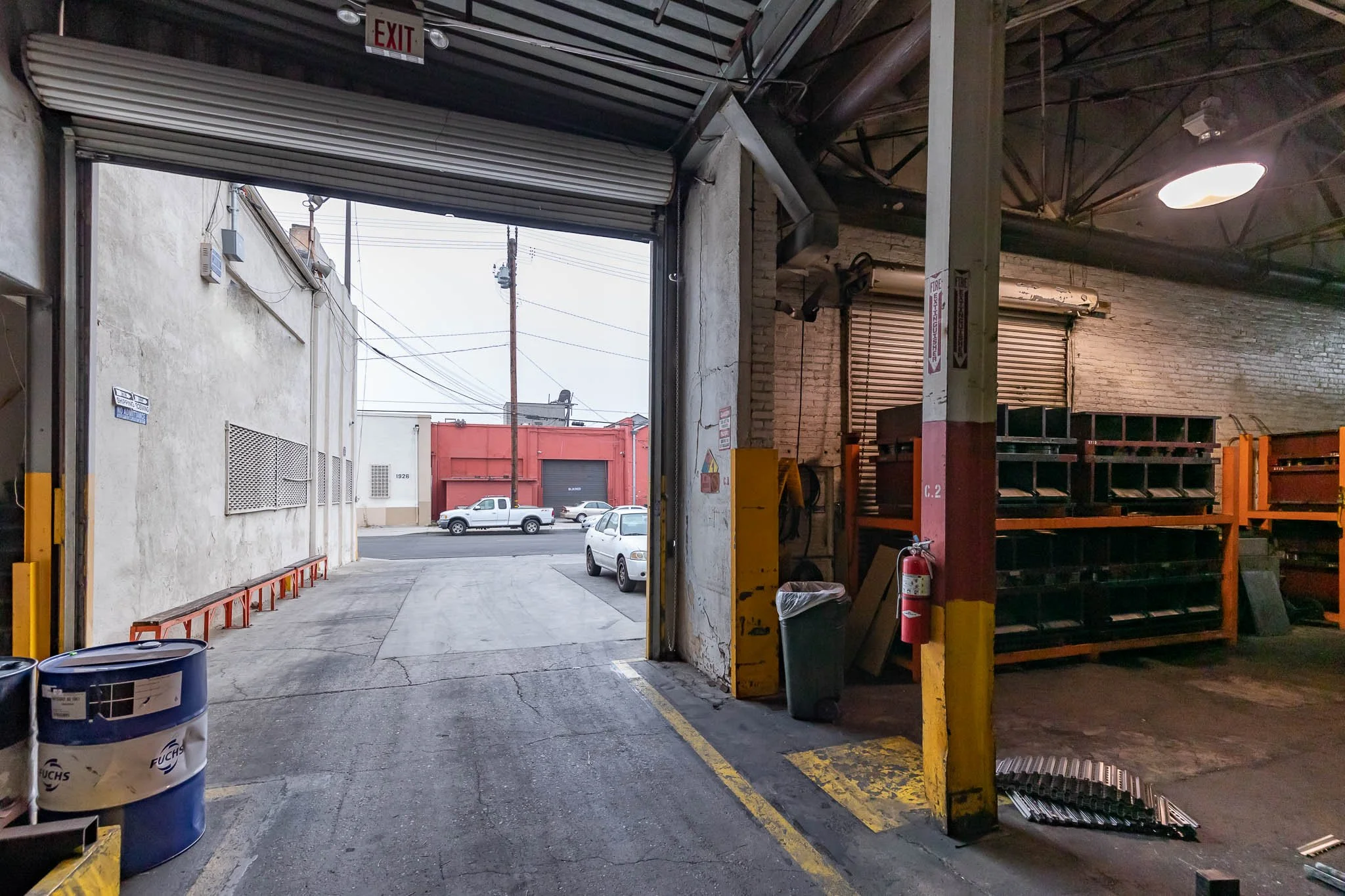 View inside a loading dock area looking outward through an open garage door to a street with parked cars and a red building in the background.