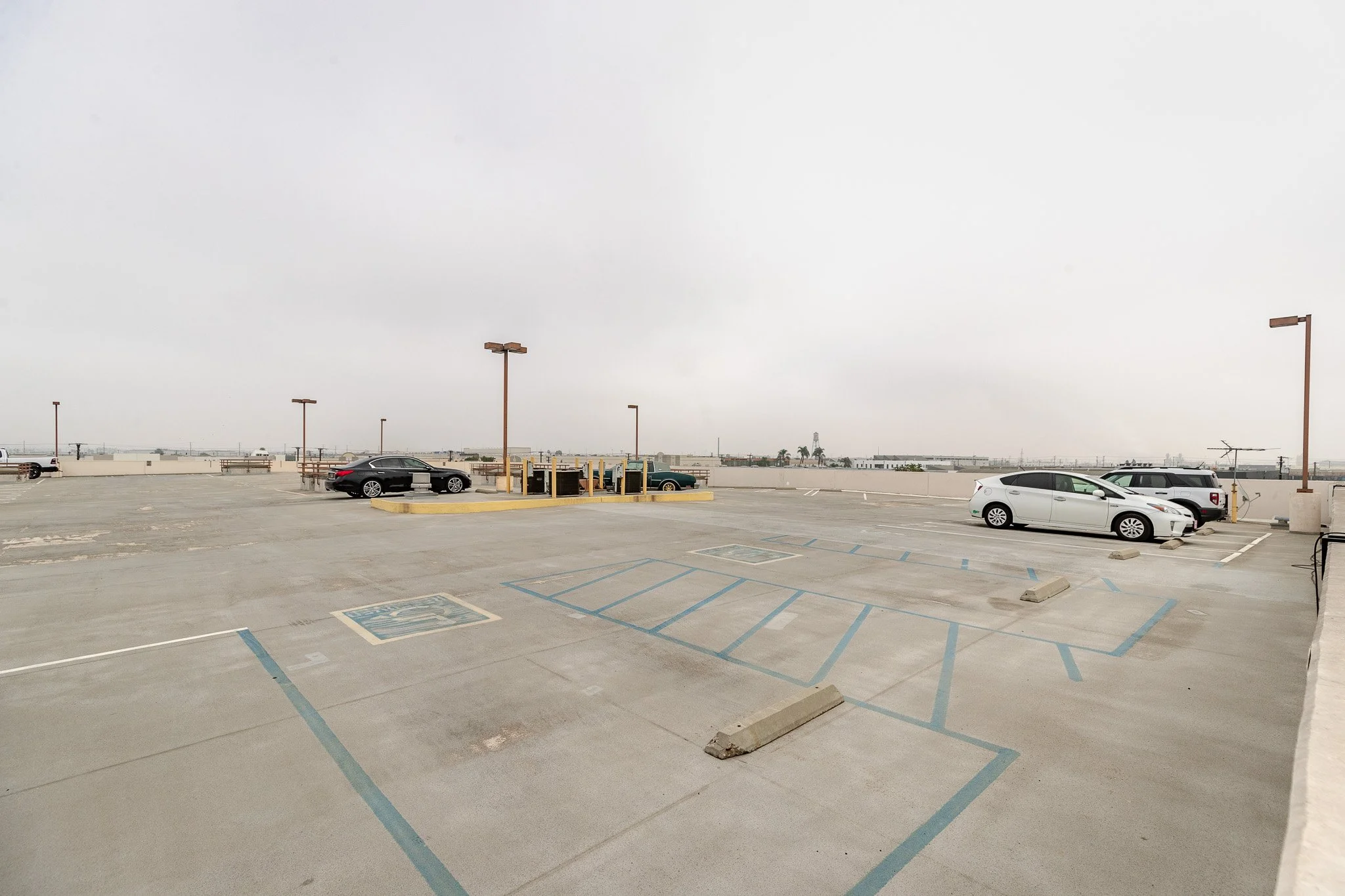Empty rooftop parking lot on a cloudy day with a few parked cars and uncovered parking spaces.