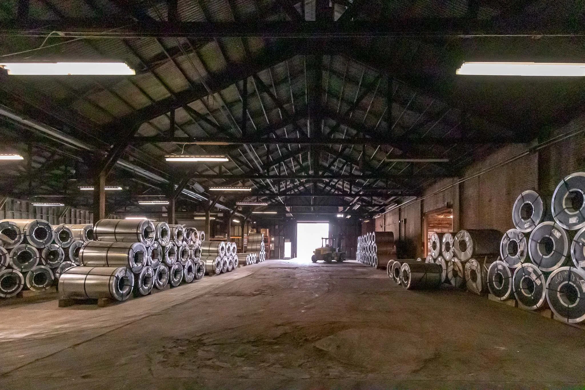 Interior of a warehouse with stacks of large metal coils on either side, a forklift in the background, and a bright light at the open doorway.