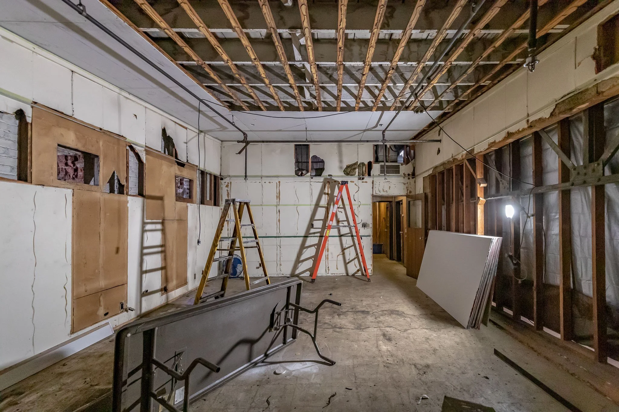 A room under renovation with exposed ceiling beams, drywall partially installed on walls, ladders, construction tools, and materials present.