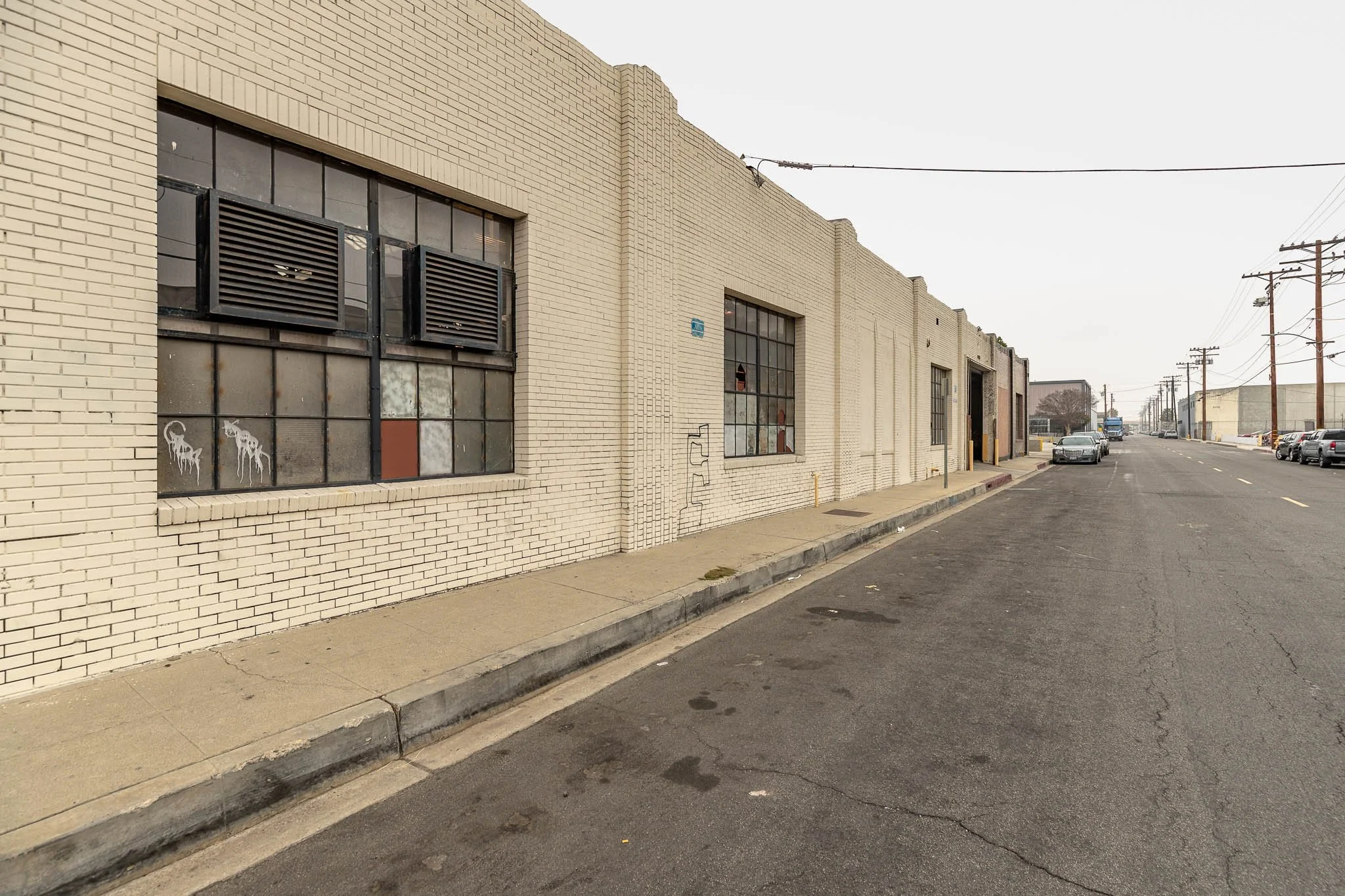 Empty city street with a beige brick building on the left, topped with large black ventilation units and graffiti, and parked cars on both sides of the street under a gray, overcast sky.