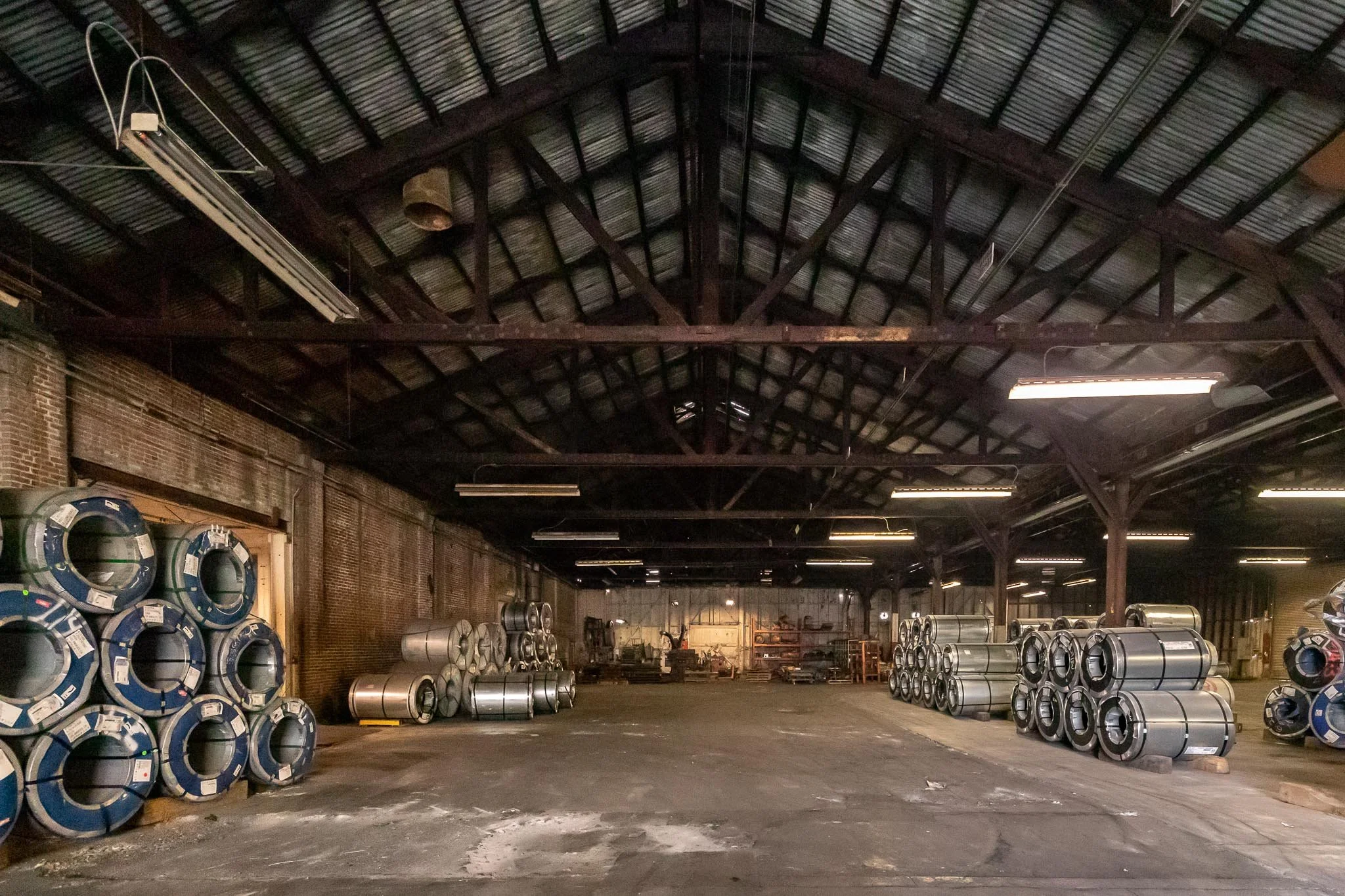 Industrial warehouse with stacked metal rolls and shelving along brick walls, illuminated by overhead fluorescent lights.