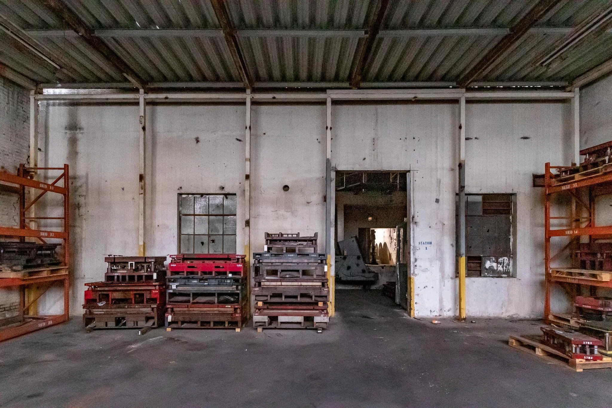 Warehouse interior with empty shelves, stacked pallets, and a doorway leading to another storage area, with a plain white wall and metal ceiling.