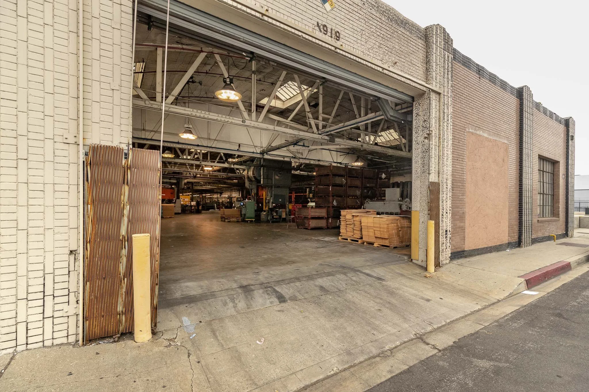The image shows an open warehouse or workshop with stacks of wood, industrial equipment, and shelving units inside. The open garage door frames the interior space.
