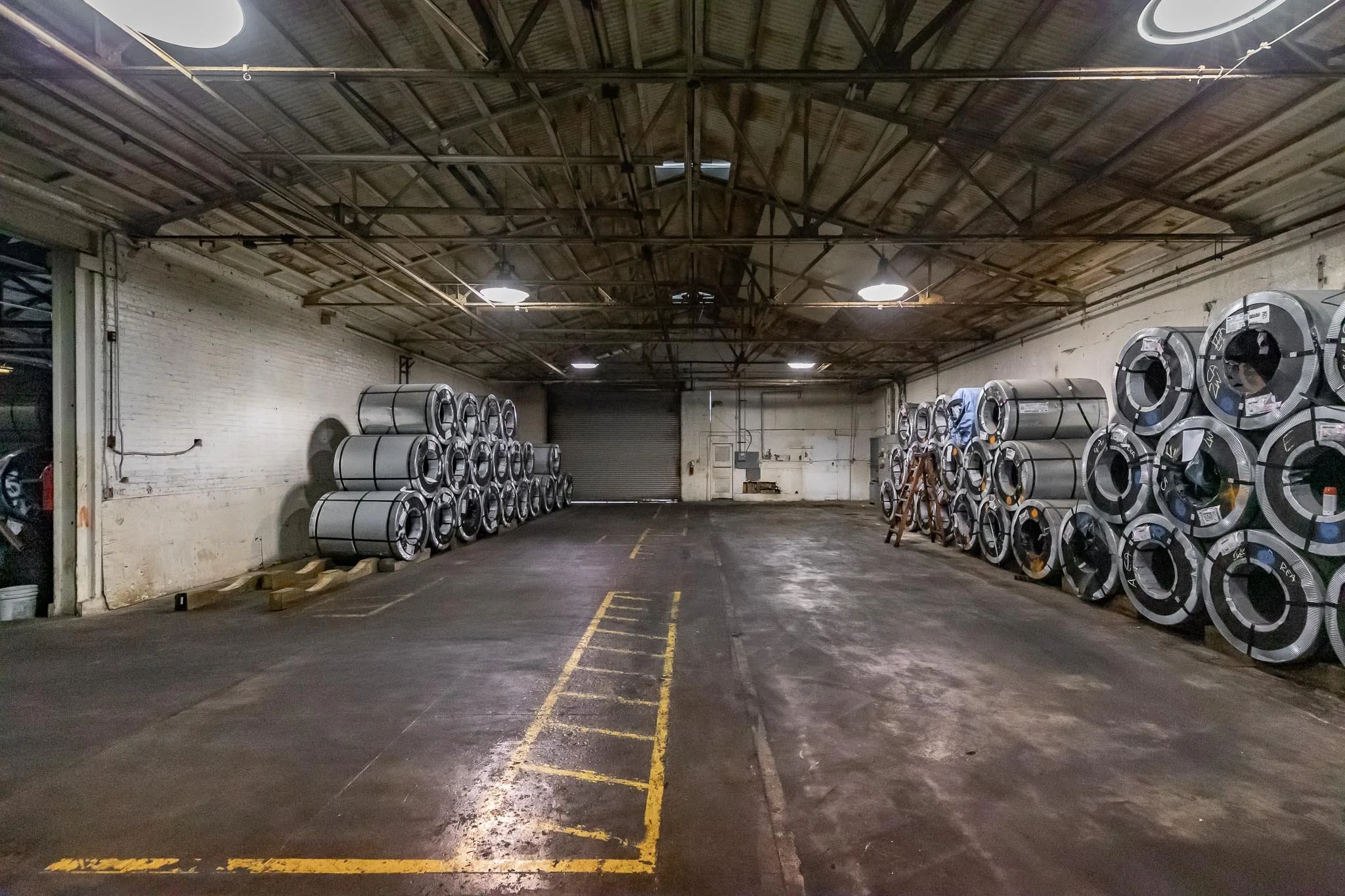 Empty industrial warehouse with stacks of metal coils on both sides and a closed garage door at the back.