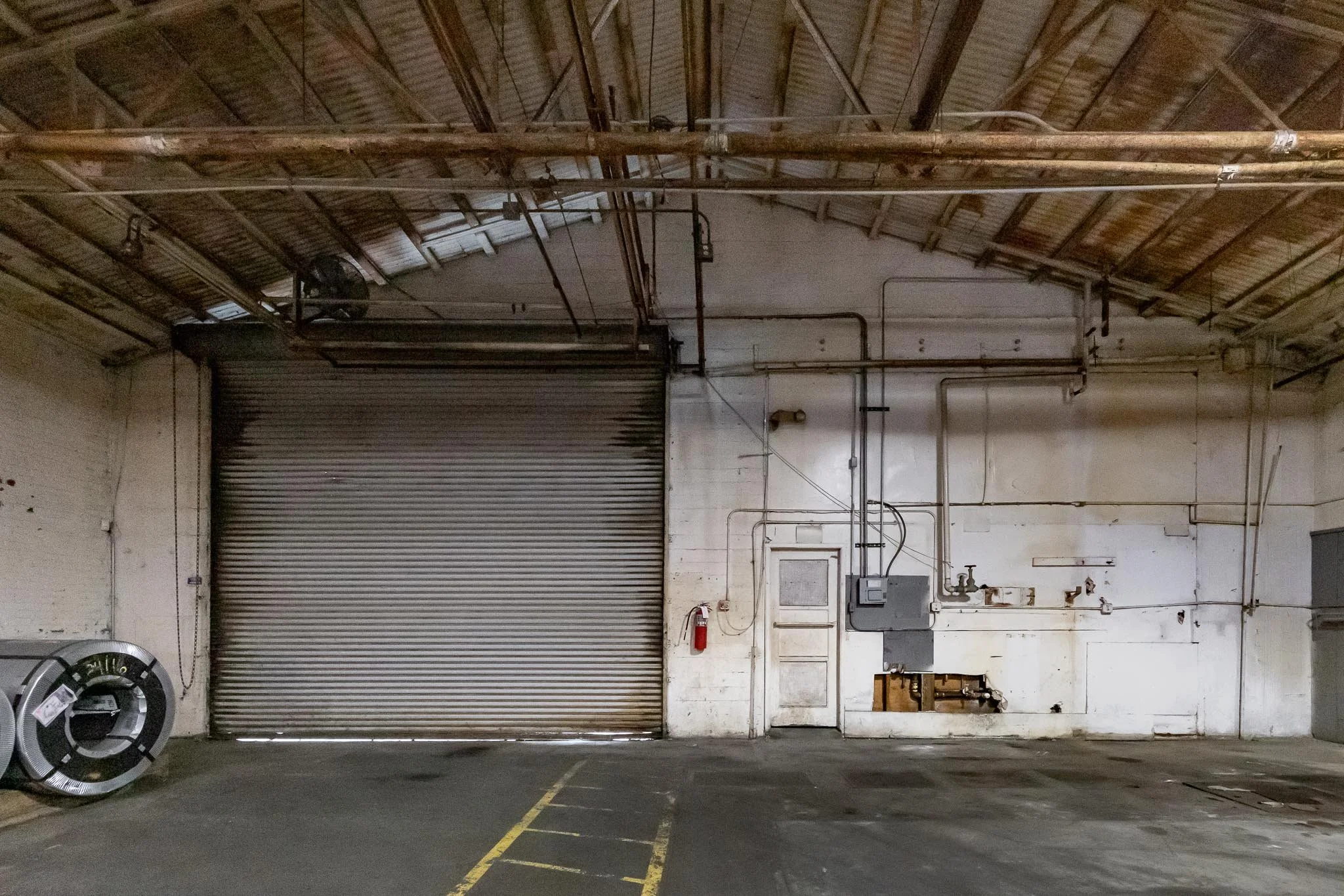 Empty warehouse with a large metal roll-up door, electrical boxes, exposed piping and wires, and a fire extinguisher on the wall.