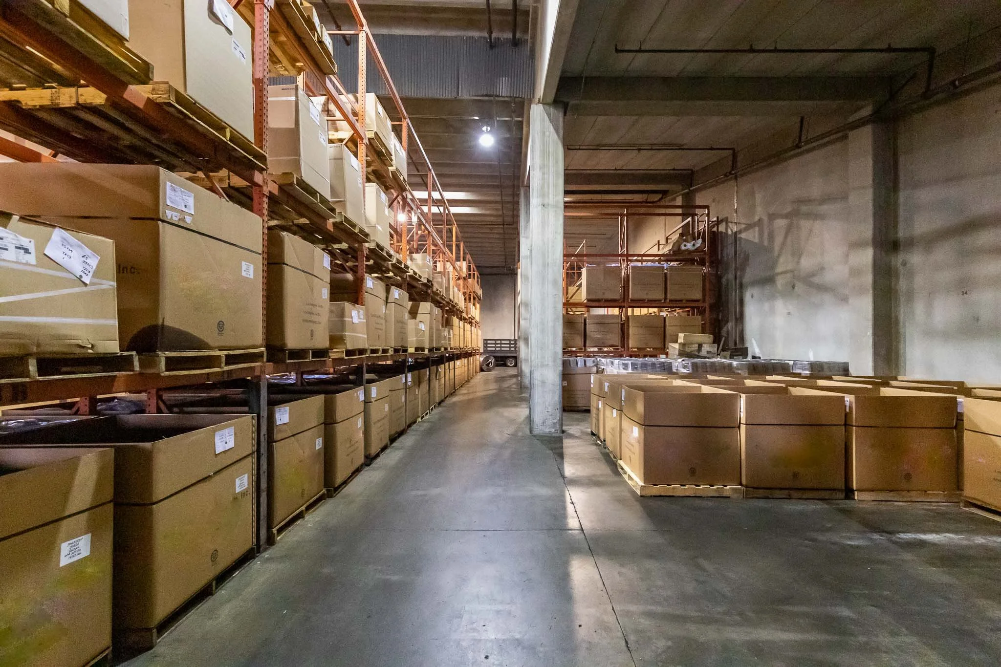 Warehouse storage area with shelves filled with cardboard boxes and pallets, concrete floor, and industrial lighting.