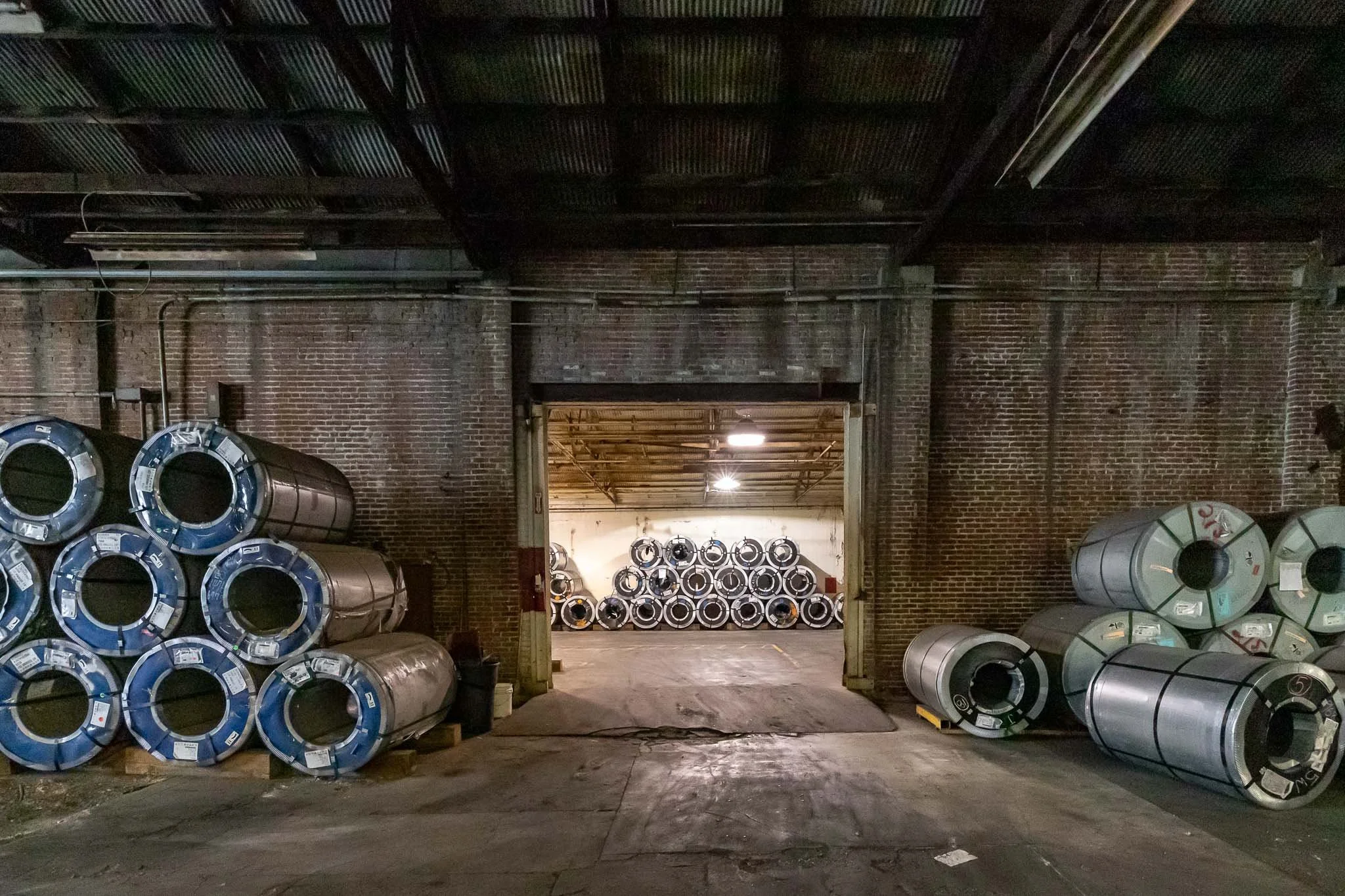 Stacked metal rolls inside an industrial warehouse with brick walls, viewed through a large garage door opening.