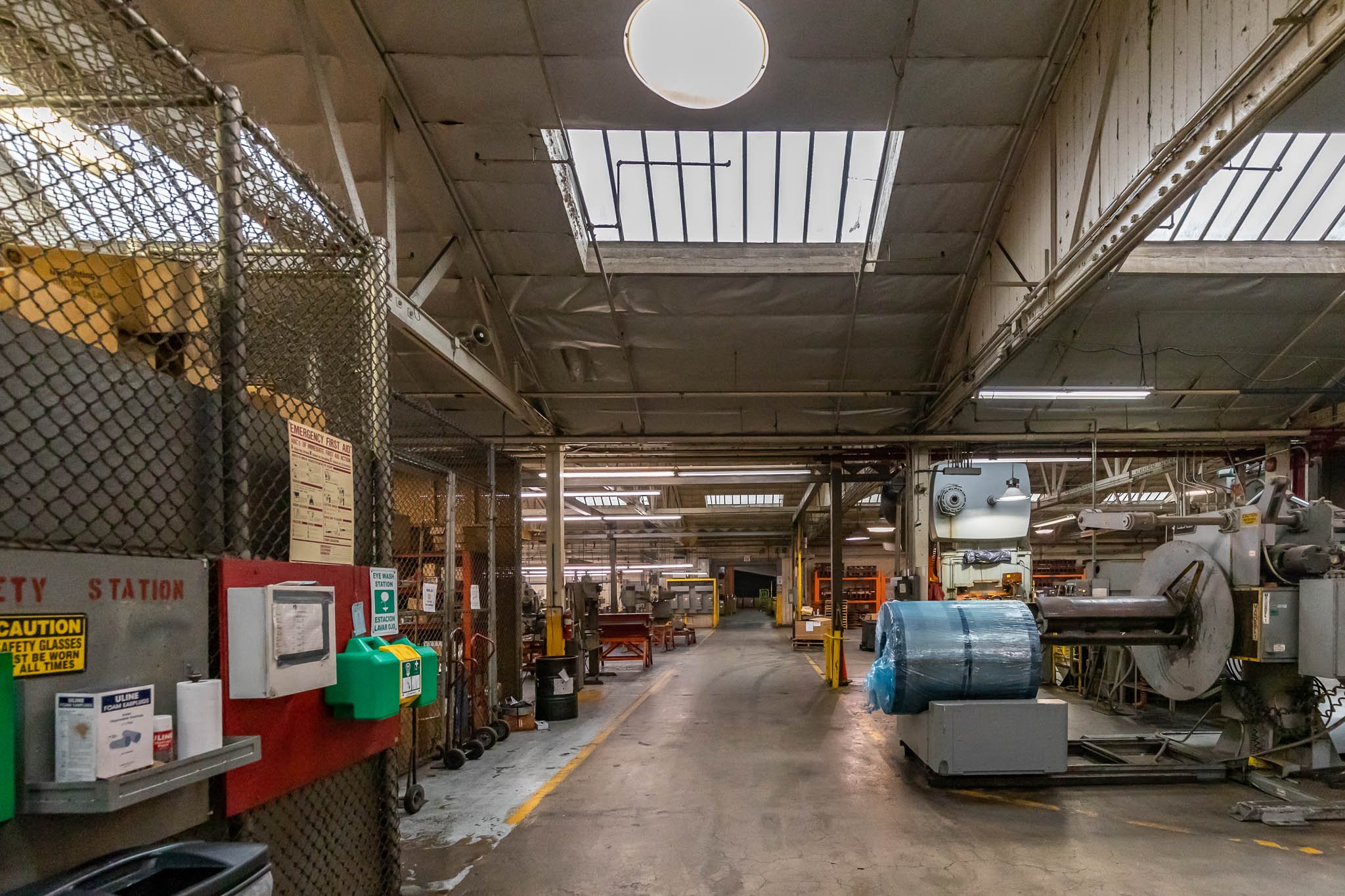 Industrial manufacturing facility with machinery, workstations, and safety equipment. The area is enclosed with a high ceiling, exposed pipes, and a skylight.