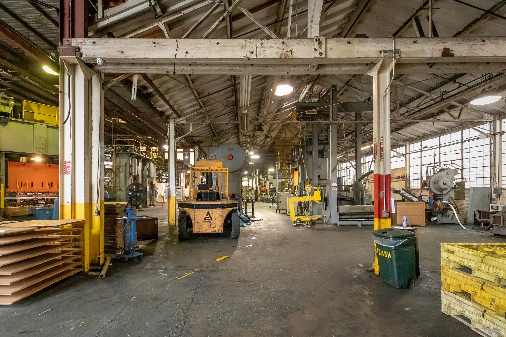 Interior of a manufacturing or workshop facility with various machines, equipment, and pallets, illuminated by overhead lights and large windows.