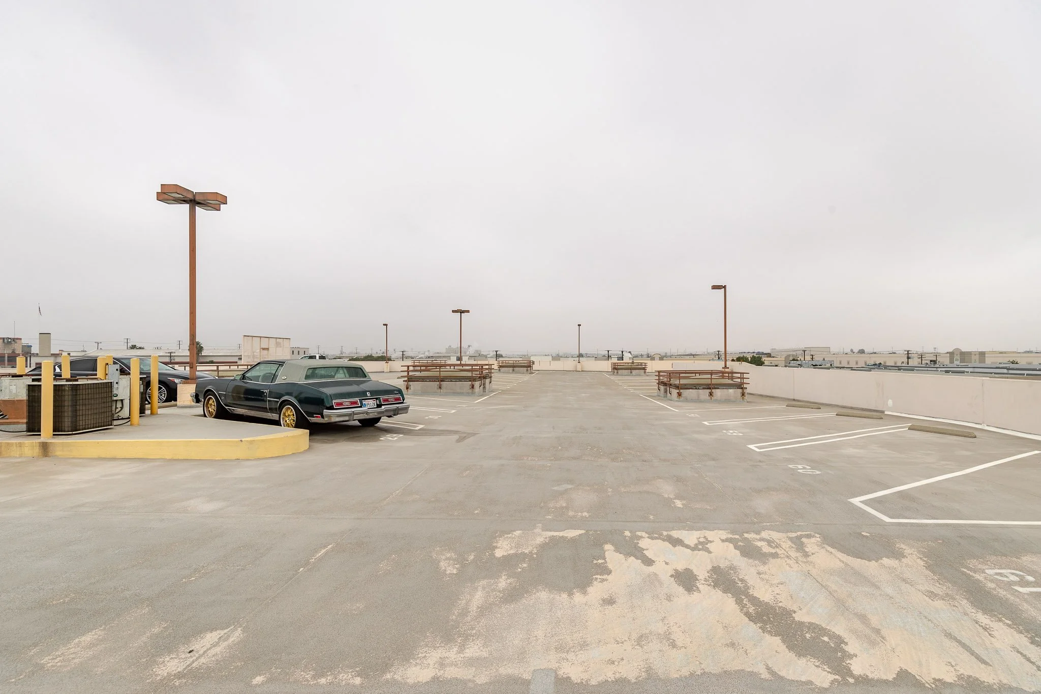 Empty rooftop parking lot with a few parked cars, lampposts, benches, and a cloudy sky.