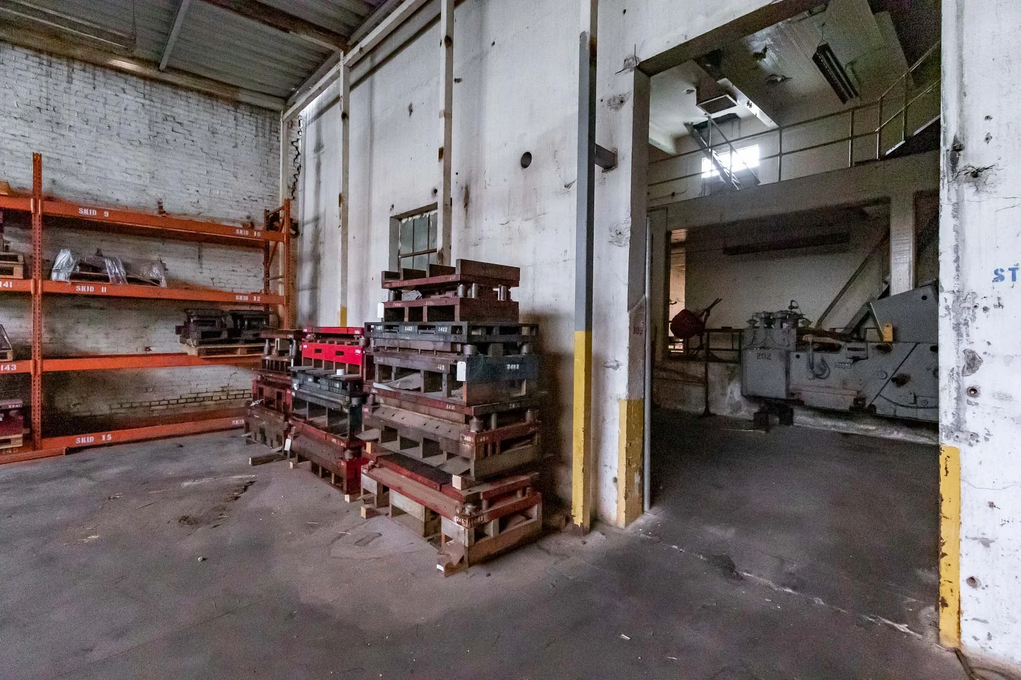Interior of an industrial warehouse with stacked pallets, metal shelving, and heavy machinery in the background.